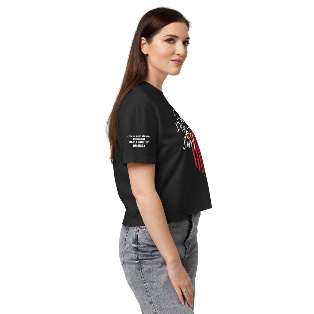 Woman wearing the black "1776 & Surf" boxy tee, side view, showing the 1776 script logo with 13-star Betsy Ross flag graphic and the 'Celebrating 250 Years of America' sleeve text – America's 250th Anniversary Collection by 17th & Surf
