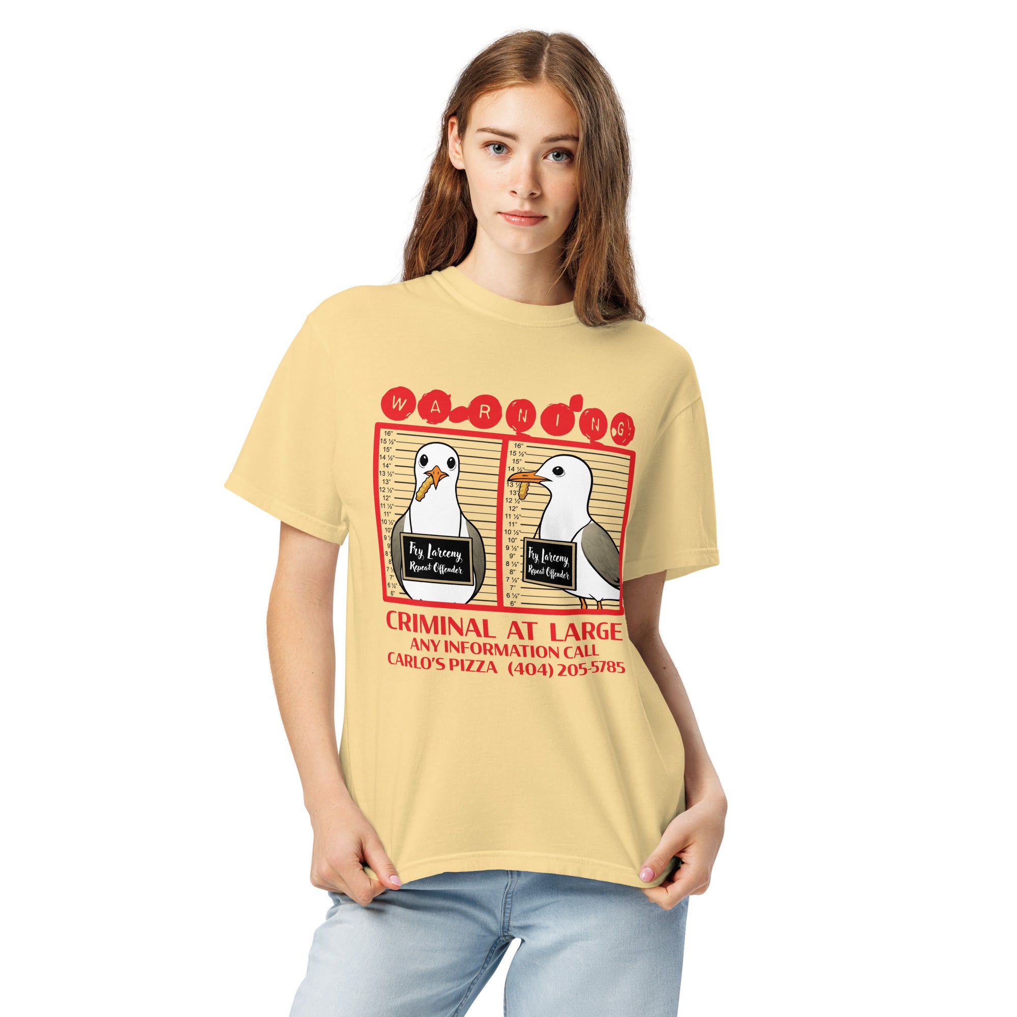 "Criminal At Large" pale yellow garment-dyed heavyweight tee on female model, front view, featuring seagull "WARNING" mugshot graphic with "Fry Larceny, Repeat Offender" police lineup – 17th & Surf x Carlo's Pizza