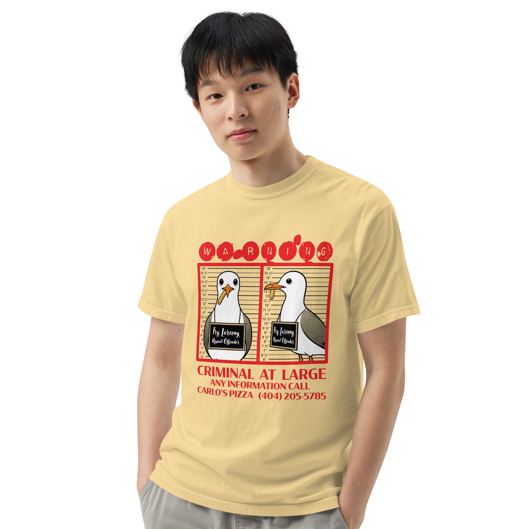 "Criminal At Large" pale yellow garment-dyed heavyweight tee on male model, front view, featuring seagull "WARNING" mugshot graphic with "Fry Larceny, Repeat Offender" police lineup – 17th & Surf x Carlo's Pizza