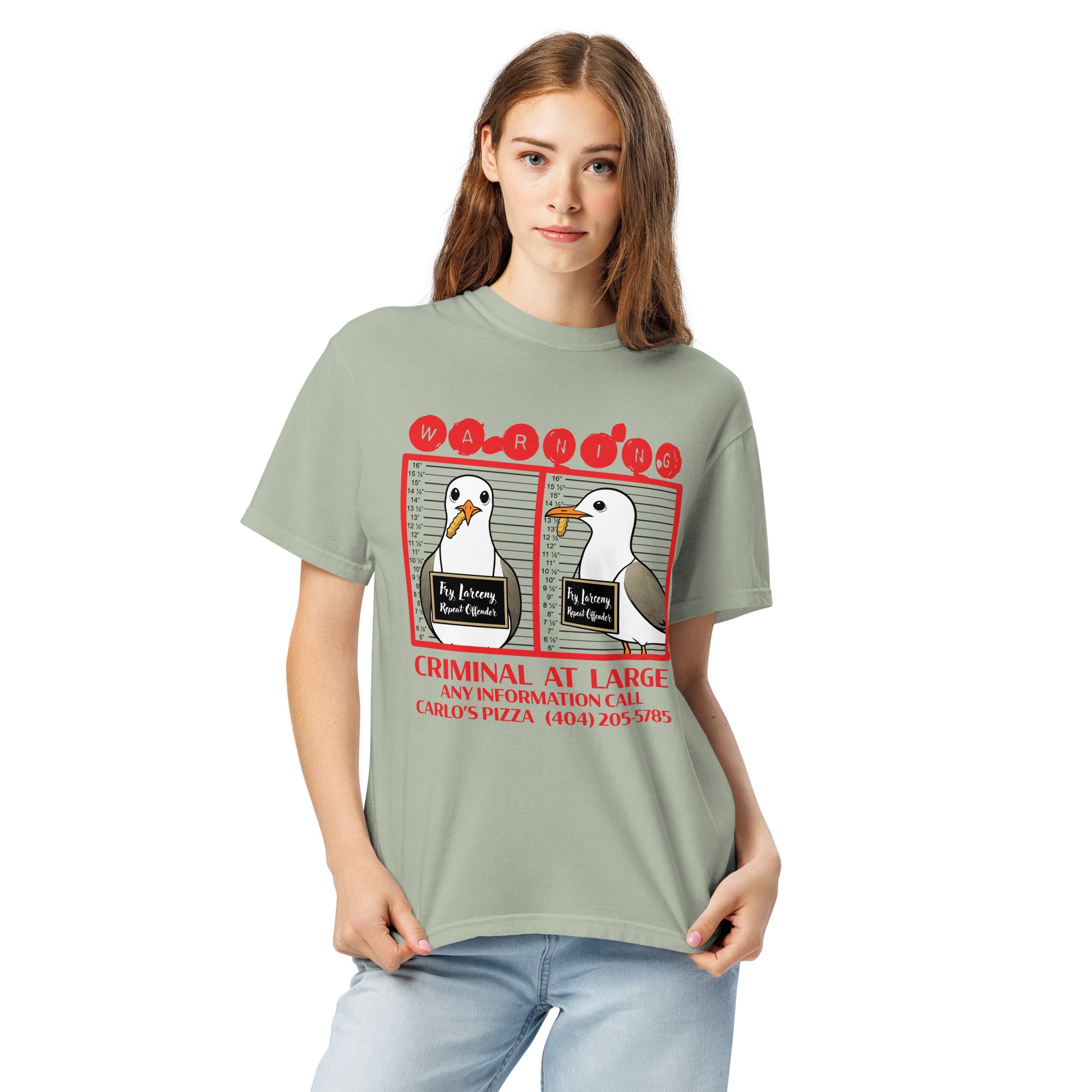 "Criminal At Large" sage green garment-dyed heavyweight tee on female model, front view, featuring seagull "WARNING" mugshot graphic with "Fry Larceny, Repeat Offender" police lineup – 17th & Surf x Carlo's Pizza