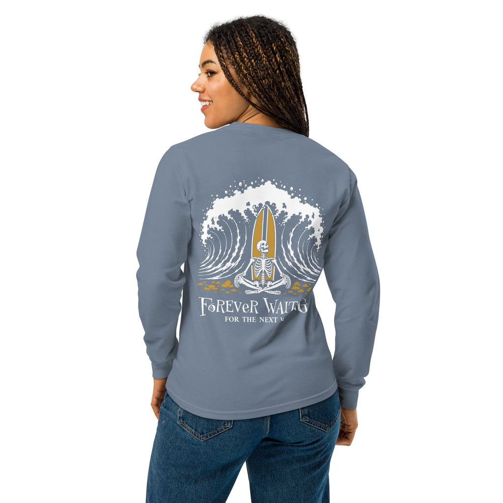 "Forever Waiting For The Next Wave" long sleeve shirt, Blue Jean, second back view, featuring a meditating skeleton surfer, board planted upright behind him, sitting cross-legged on the shore as breakers roll in on either side – 17th & Surf