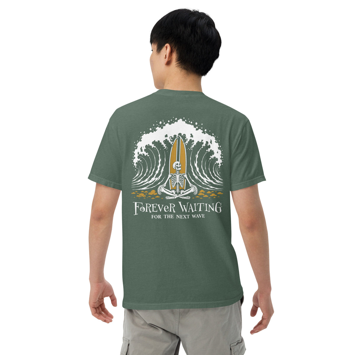 "Forever Waiting For The Next Wave" tee, Blue Spruce, fourth back view, featuring a meditating skeleton surfer, board planted upright behind him, sitting cross-legged on the shore as breakers roll in on either side – 17th & Surf
