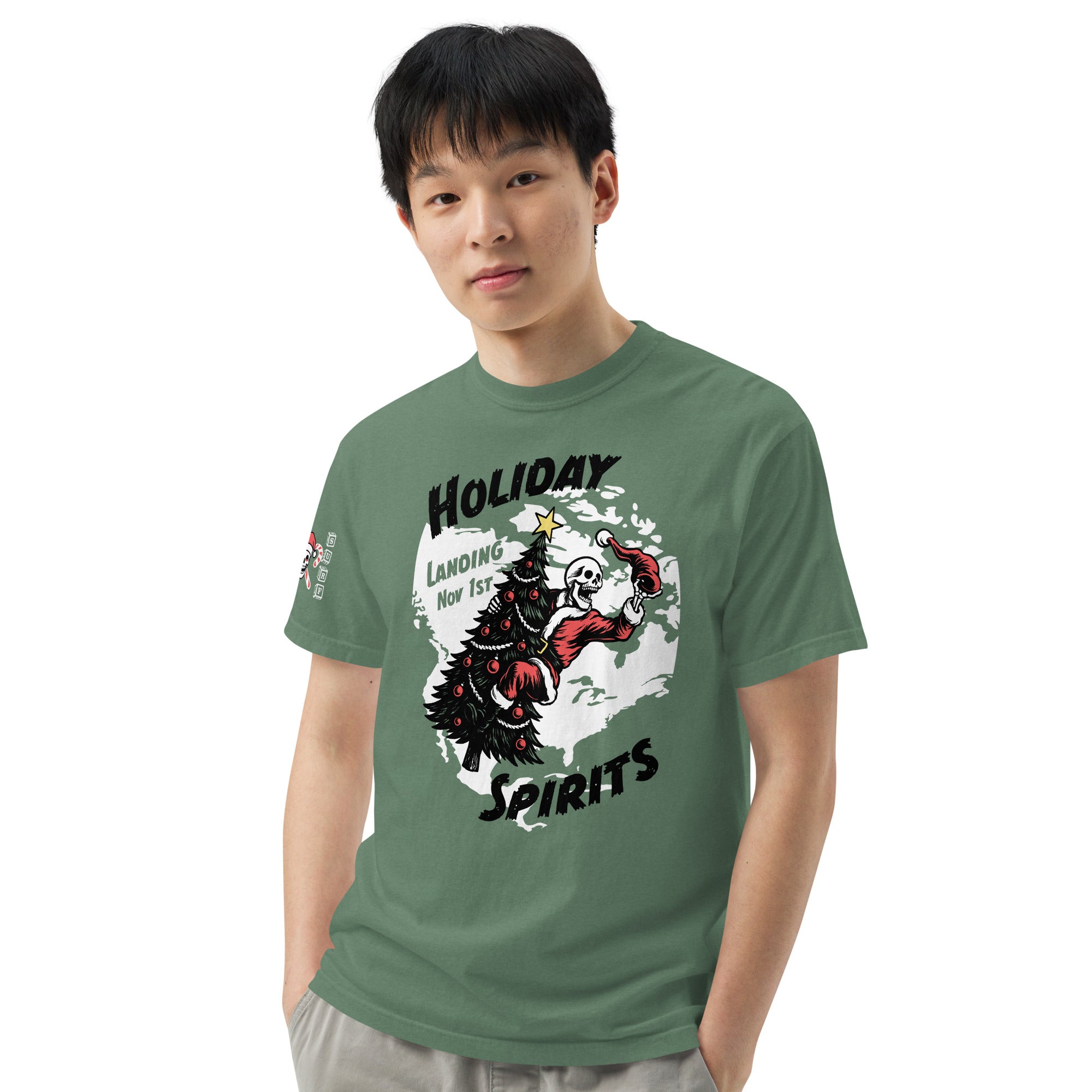 Model wearing "Holiday Spirits" front print garment-dyed heavyweight t-shirt in light green, third front view featuring full color graphic of a skeleton Santa riding a decorated Christmas tree inside a white splatter map shape with arched "Holiday" and "Spirits" text and "Landing Nov 1st" – 17th & Surf