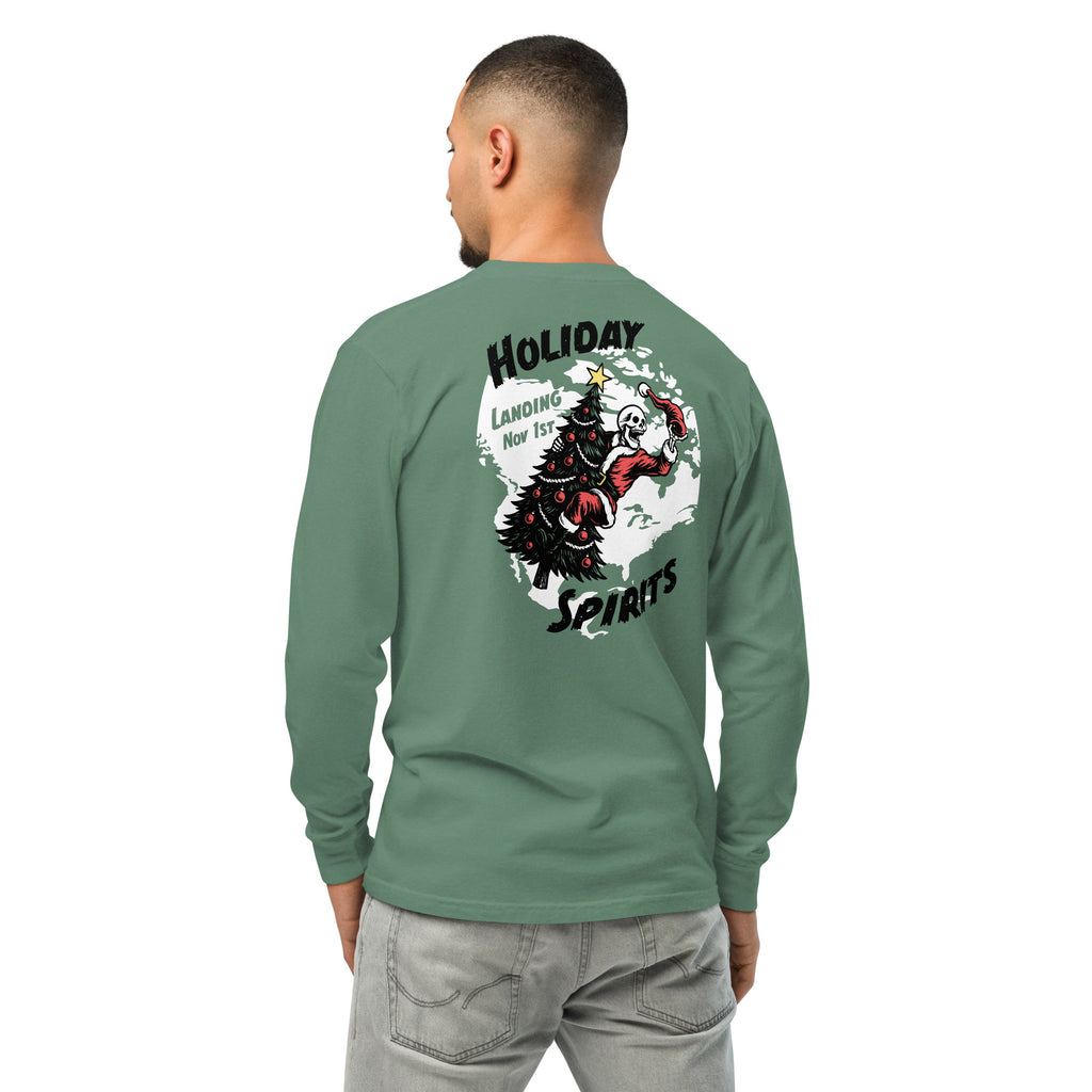 Model wearing "Holiday Spirits" garment-dyed heavyweight long sleeve shirt in sage green, second back angle view featuring full color graphic of a skeleton Santa riding a decorated Christmas tree inside a white splatter map shape with arched "Holiday" and "Spirits" text and "Landing Nov 1st" – 17th & Surf