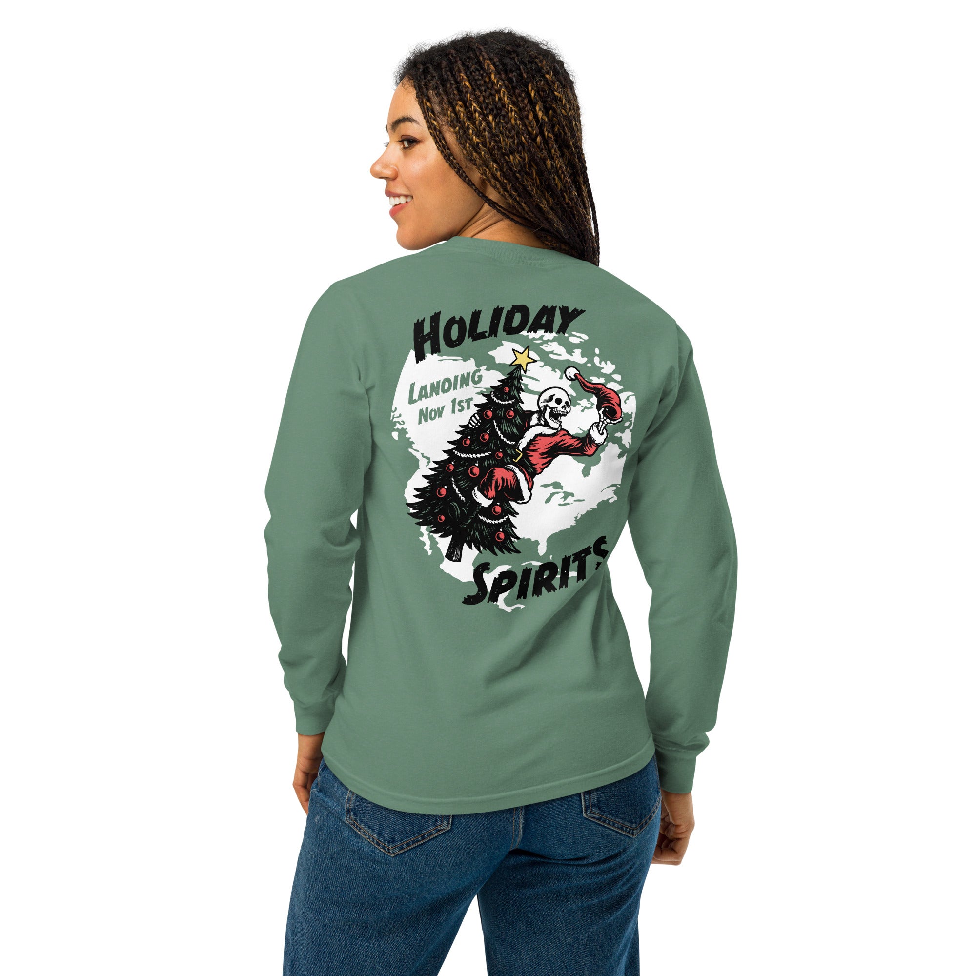 Model wearing "Holiday Spirits" garment-dyed heavyweight long sleeve shirt in sage green, back angle view featuring full color graphic of a skeleton Santa riding a decorated Christmas tree inside a white splatter map shape with arched "Holiday" and "Spirits" text and "Landing Nov 1st" – 17th & Surf