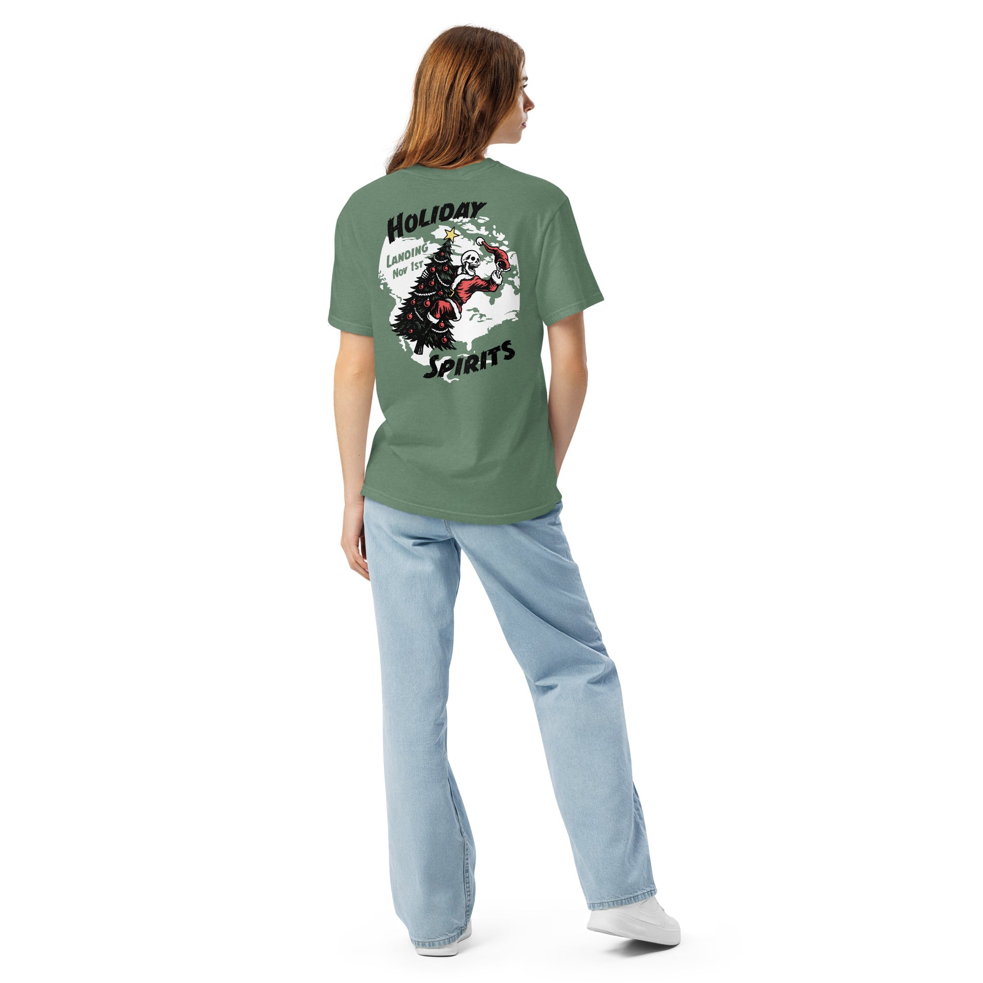 Model wearing "Holiday Spirits" garment-dyed heavyweight t-shirt in light green, second back angle view featuring full color graphic of a skeleton Santa riding a decorated Christmas tree inside a white splatter map shape with arched "Holiday" and "Spirits" text and "Landing Nov 1st" – 17th & Surf