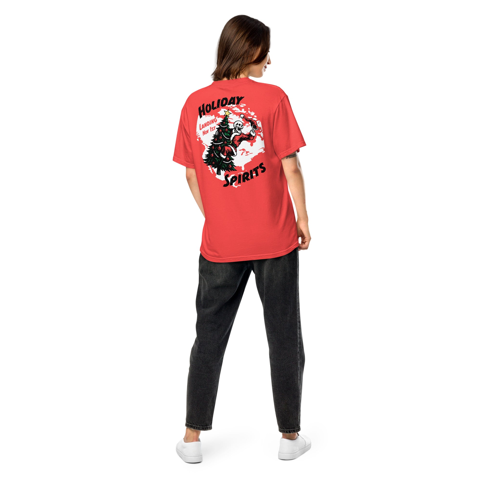 Model wearing "Holiday Spirits" garment-dyed heavyweight t-shirt in red, back angle view featuring full color graphic of a skeleton Santa riding a decorated Christmas tree inside a white splatter map shape with arched "Holiday" and "Spirits" text and "Landing Nov 1st" – 17th & Surf