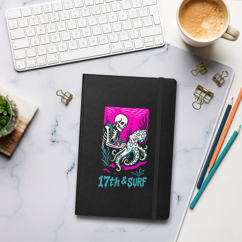 "Inked" hardcover notebook lifestyle flat lay on desk in black featuring skeleton tattooing an octopus graphic with "17th & Surf" text – 17th & Surf