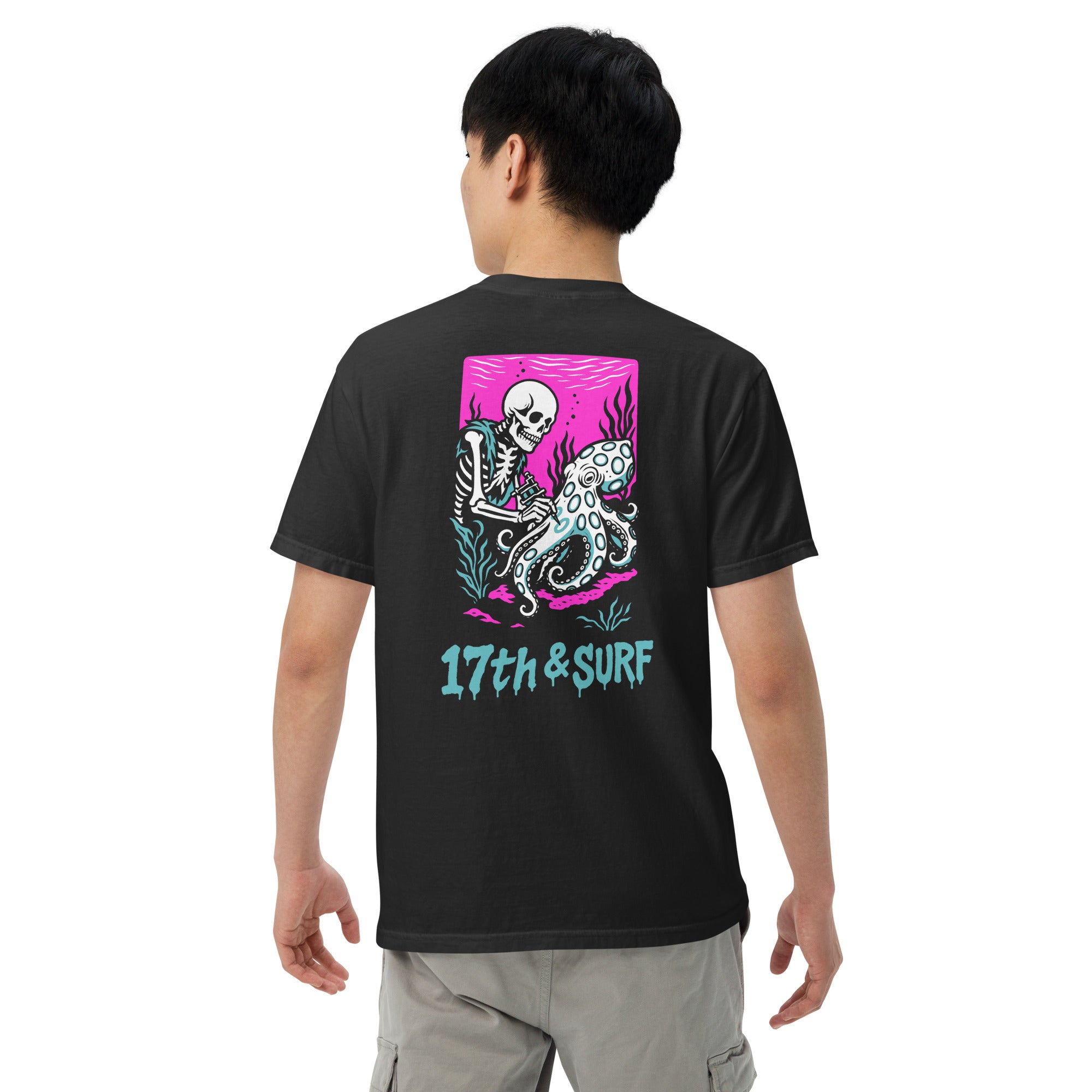 "Inked" garment-dyed heavyweight tee back view on male model in black featuring skeleton tattooing an octopus graphic with "17th & Surf" text on pink background – 17th & Surf