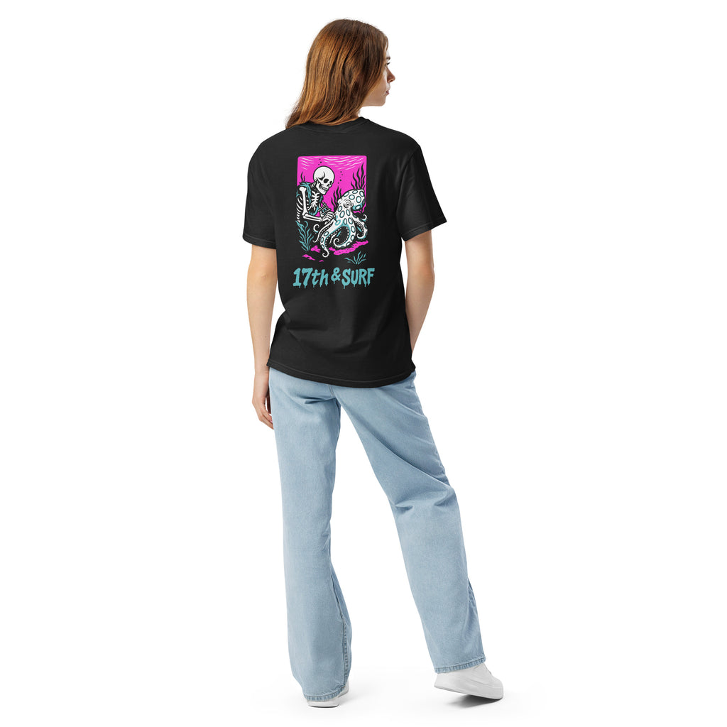 "Inked" garment-dyed heavyweight tee back view on female model in black featuring skeleton tattooing an octopus graphic with "17th & Surf" text on pink background – 17th & Surf