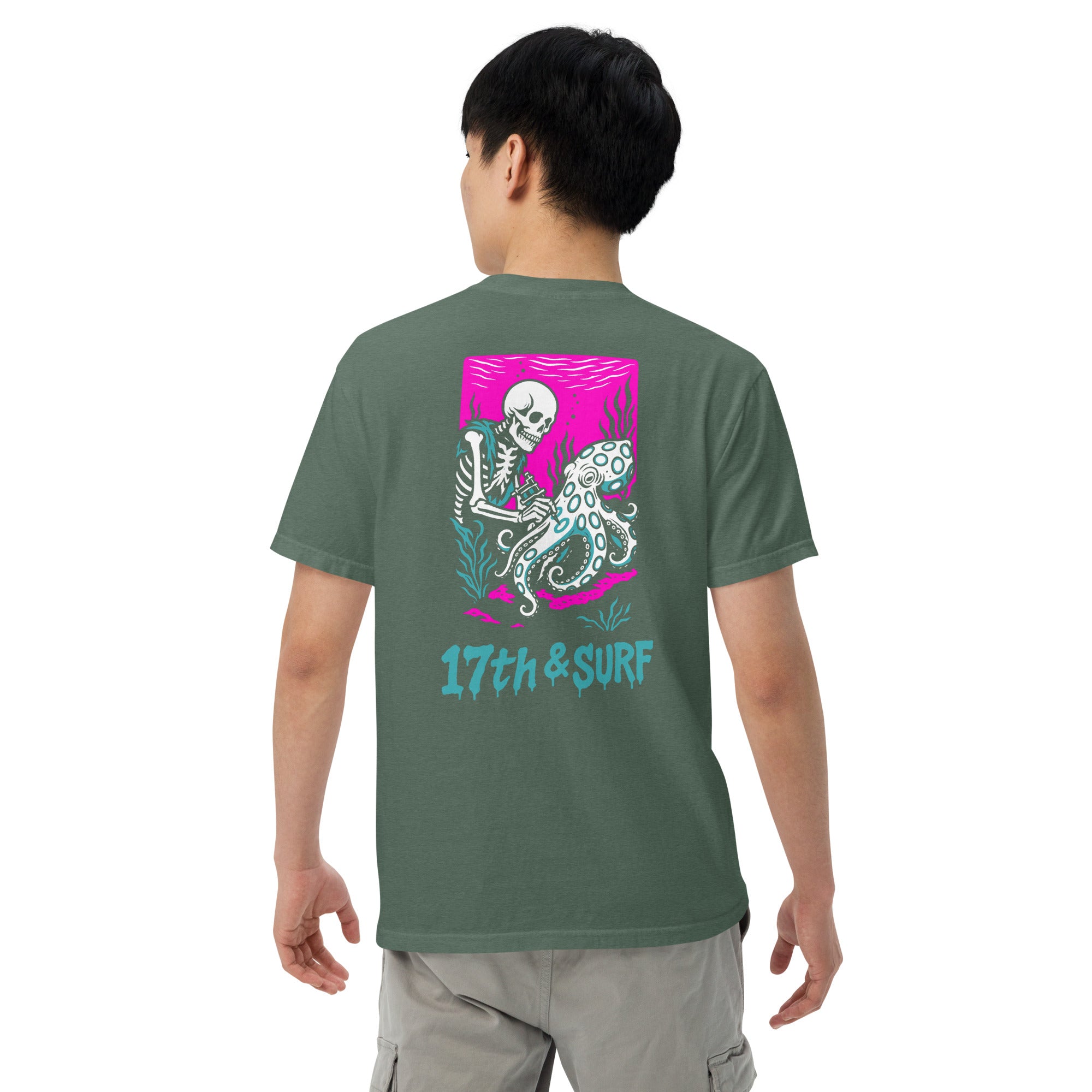 "Inked" garment-dyed heavyweight tee back angle in blue spruce featuring skeleton tattooing an octopus graphic with "17th & Surf" text on pink background – 17th & Surf