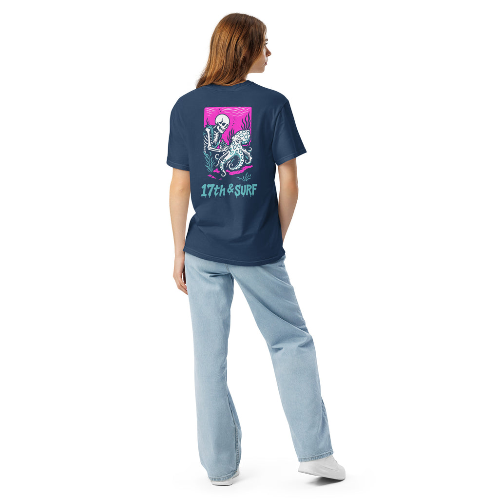 "Inked" garment-dyed heavyweight tee back view on female model in true navy featuring skeleton tattooing an octopus graphic with "17th & Surf" text on pink background – 17th & Surf