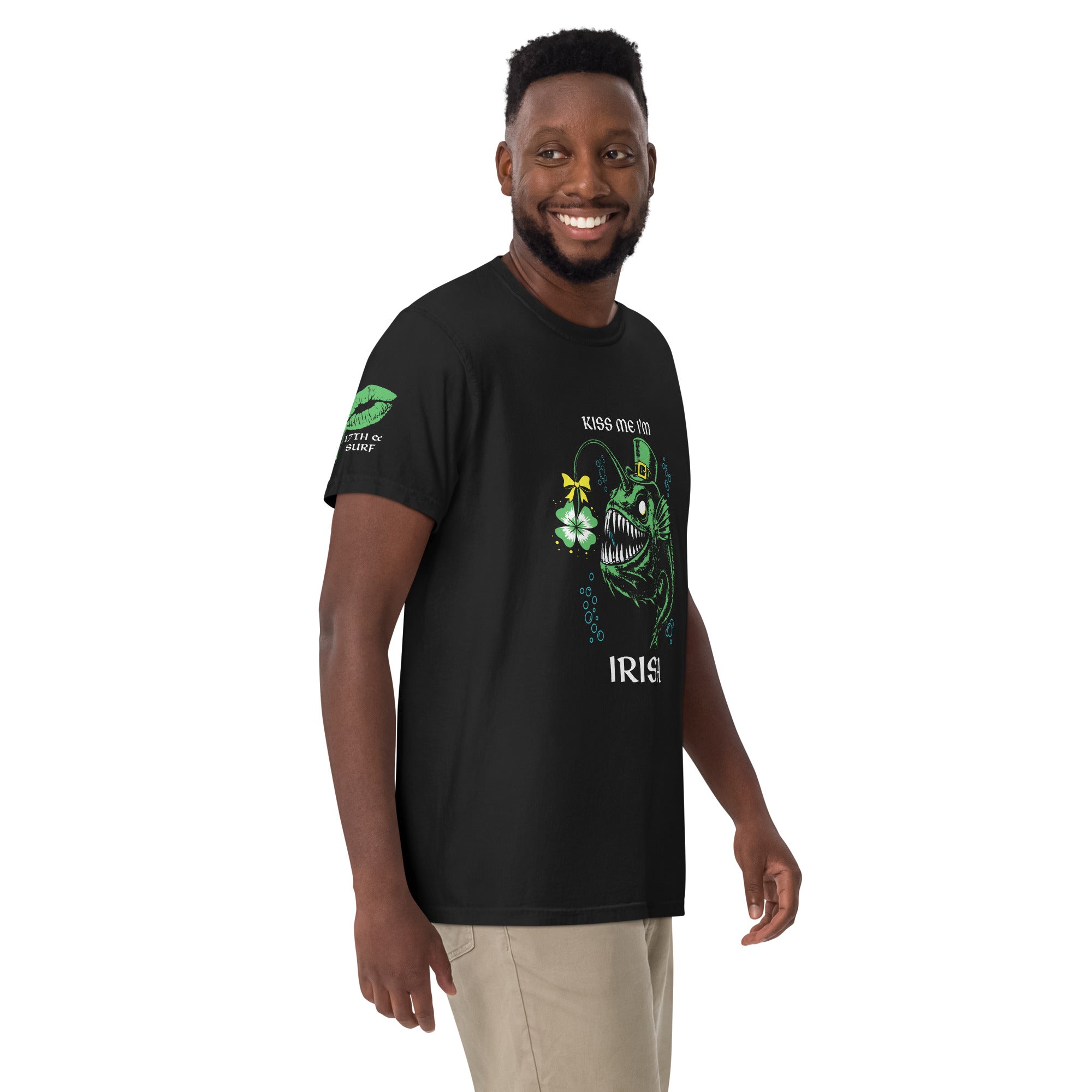 "Kiss Me I'm Irish" Anglerfish black tee on male model, front view, featuring the green leprechaun anglerfish with shamrock lure and "KISS ME I'M IRISH" text, plus a dripping-lips "17TH & SURF" sleeve logo – 17th & Surf