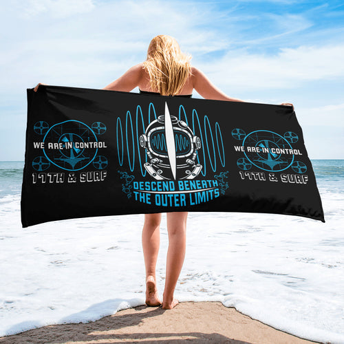 "Beneath The Outer Limits" white 30x60 beach towel, held by woman on the beach, featuring the large center graphic of a vintage diving helmet split by a surfboard fin with the text "DESCEND BENEATH THE OUTER LIMITS," flanked by two small blue "WE ARE IN CONTROL / 17th & Surf" broadcast test-pattern logos – "Beneath The Outer Limits" Series by 17th & Surf