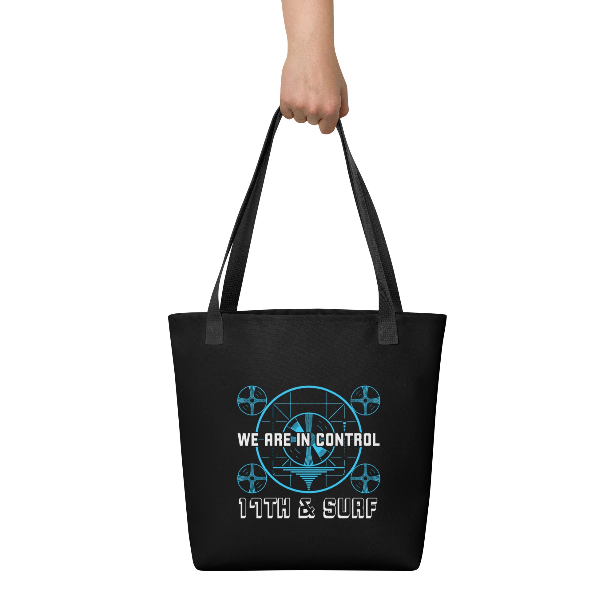 "Beneath The Outer Limits" black all-over print tote bag, back view held by model, featuring the small blue "WE ARE IN CONTROL" broadcast test-pattern logo with "17th & Surf" text – "Beneath The Outer Limits" Series by 17th & Surf