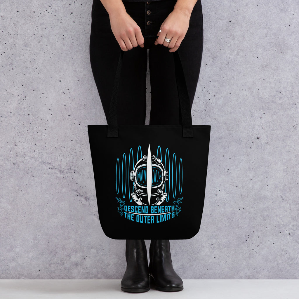 "Beneath The Outer Limits" black all-over print tote bag, front view on female model, featuring the large graphic of a vintage diving helmet split by a surfboard fin, framed by blue broadcast test-pattern rings with an oscillating waveform and the text "DESCEND BENEATH THE OUTER LIMITS" – "Beneath The Outer Limits" Series by 17th & Surf