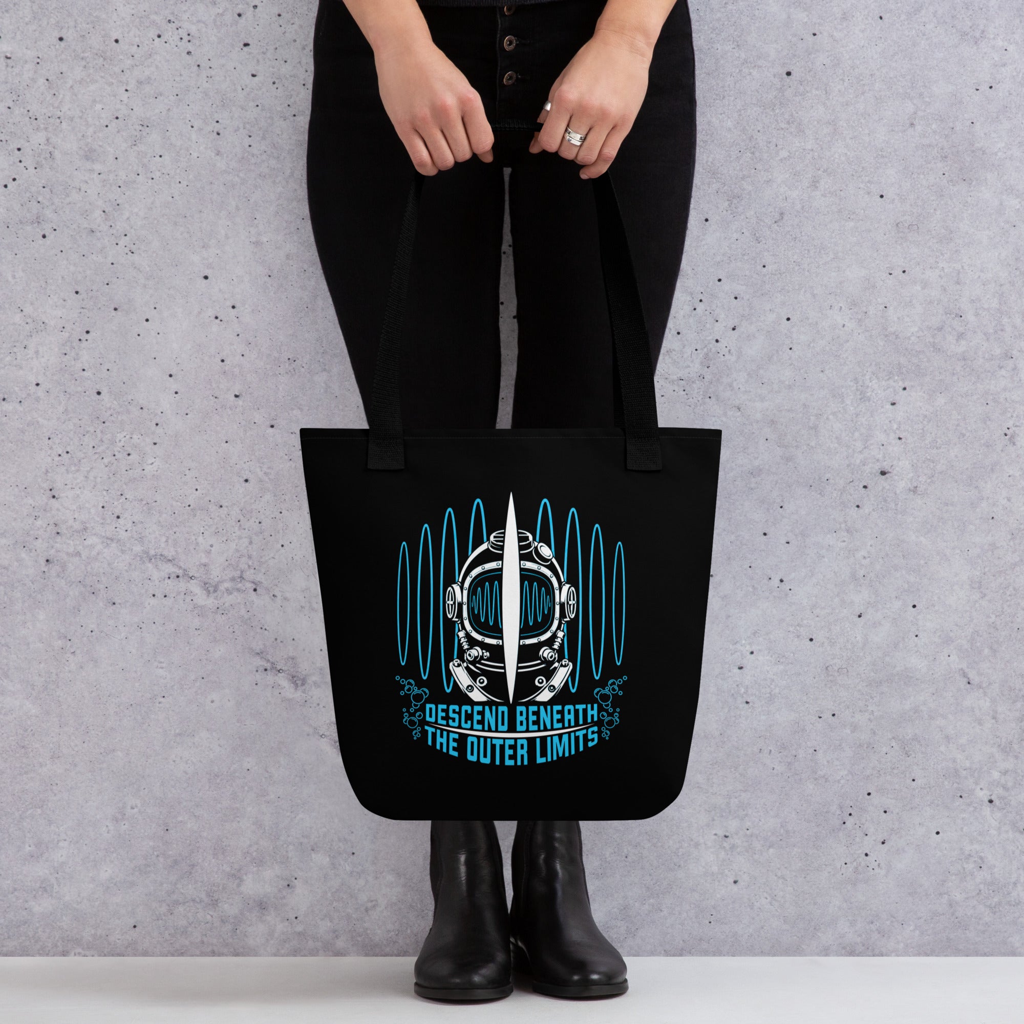 "Beneath The Outer Limits" black all-over print tote bag, front view on female model, featuring the large graphic of a vintage diving helmet split by a surfboard fin, framed by blue broadcast test-pattern rings with an oscillating waveform and the text "DESCEND BENEATH THE OUTER LIMITS" – "Beneath The Outer Limits" Series by 17th & Surf