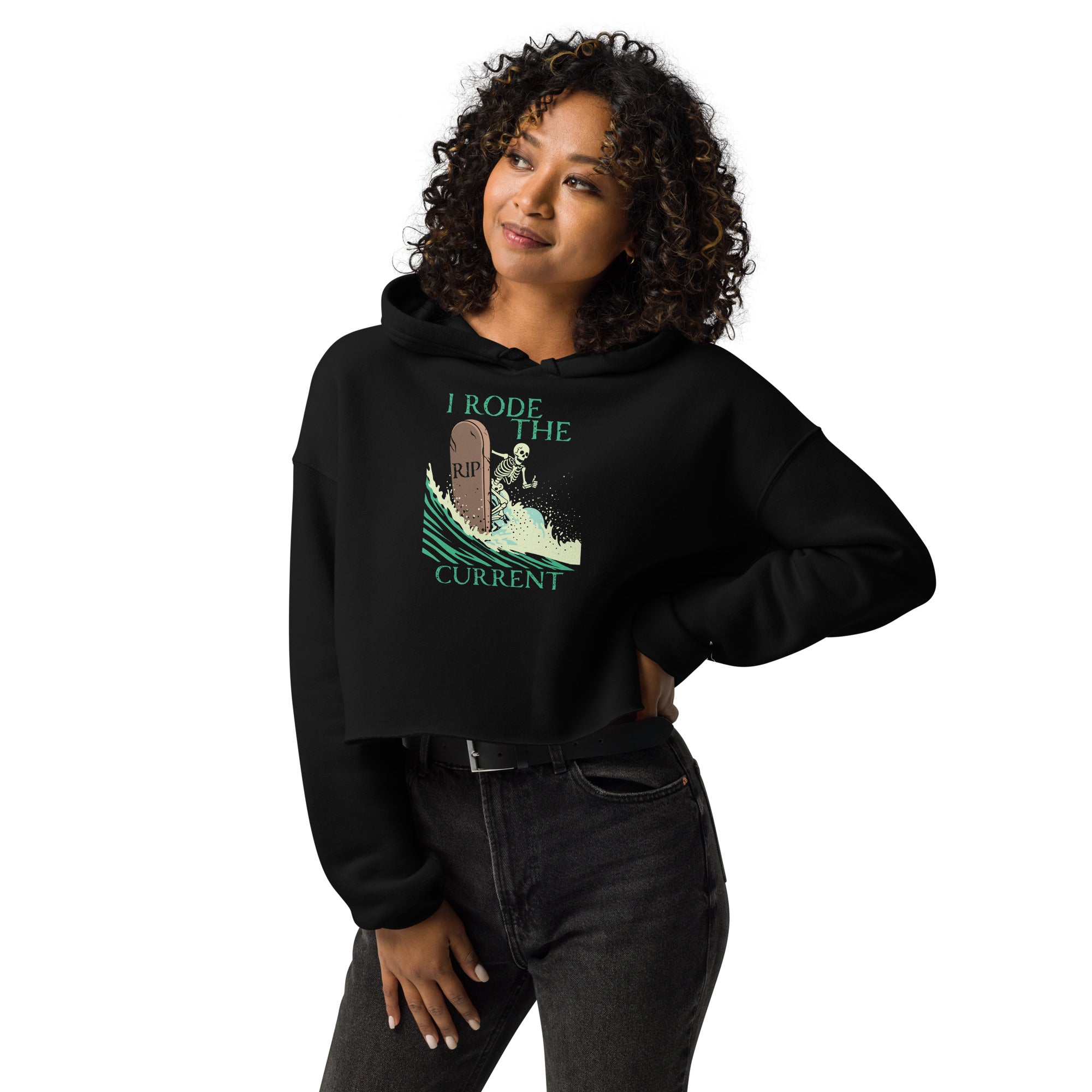 Model wearing "RIP Current" women's crop hoodie in black featuring skeleton surfing a "RIP" tombstone "I Rode the Current" graphic – 17th & Surf