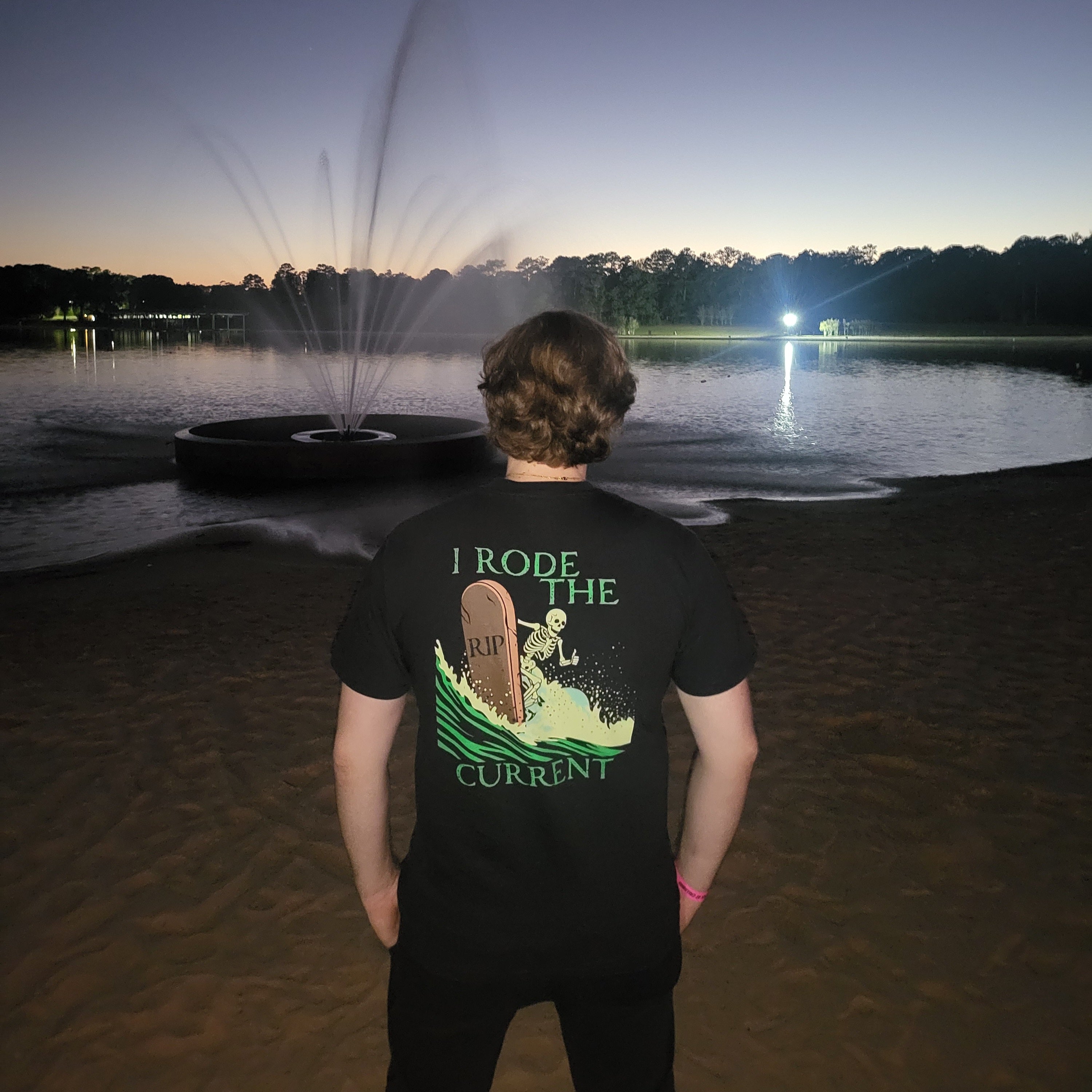 Lifestyle photo of "RIP Current" black tee with skeleton surfing a "RIP" tombstone "I Rode the Current" graphic, worn outdoors at dusk – 17th & Surf