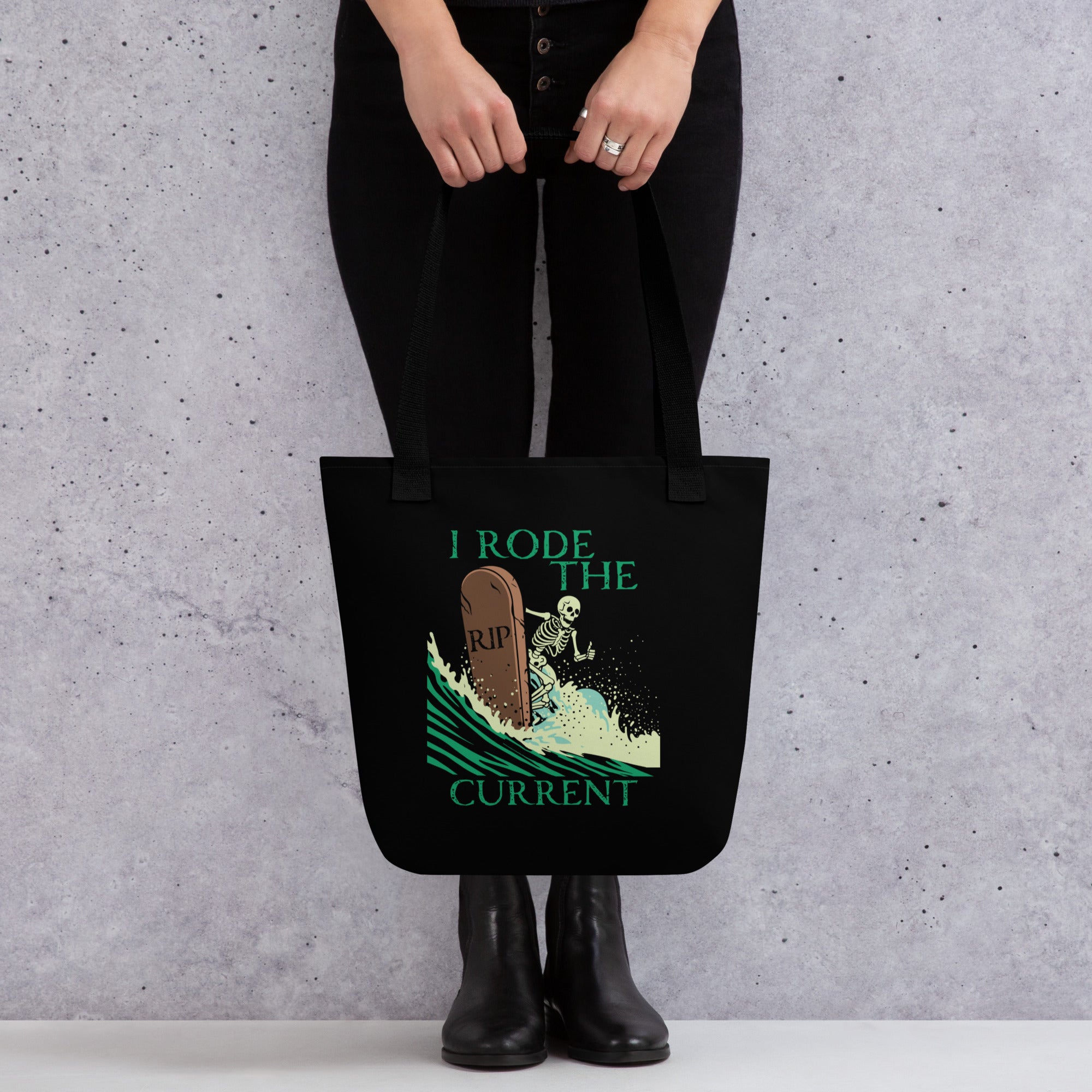 Model holding "RIP Current" all-over print tote bag in black featuring skeleton surfing a "RIP" tombstone "I Rode the Current" graphic – 17th & Surf