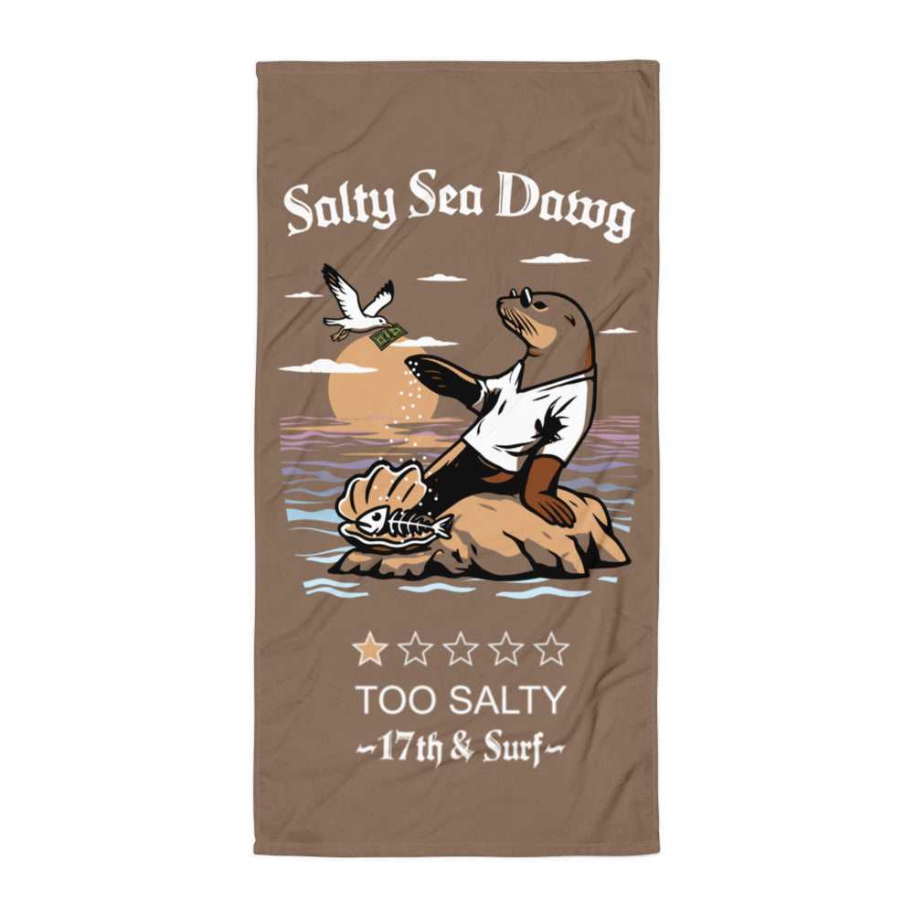 "Salty Sea Dawg" beach towel flat lay featuring color graphic of a sea lion sprinkling salt on a clam shell with a fish skeleton beneath arched "Salty Sea Dawg" text and "TOO SALTY" one-star rating with "17th & Surf" wordmark – 17th & Surf