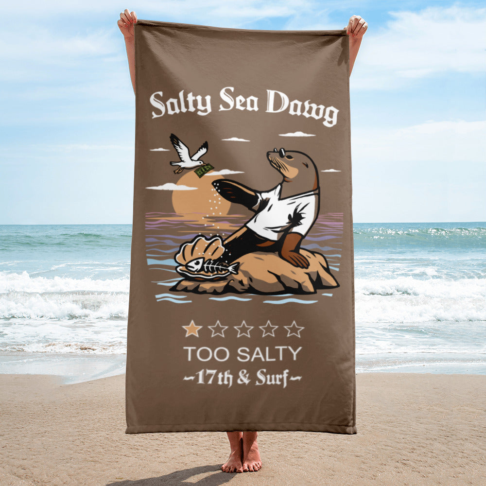 "Salty Sea Dawg" beach towel held up on the beach featuring color graphic of a sea lion sprinkling salt on a clam shell with a fish skeleton beneath arched "Salty Sea Dawg" text and "TOO SALTY" one-star rating with "17th & Surf" wordmark – 17th & Surf