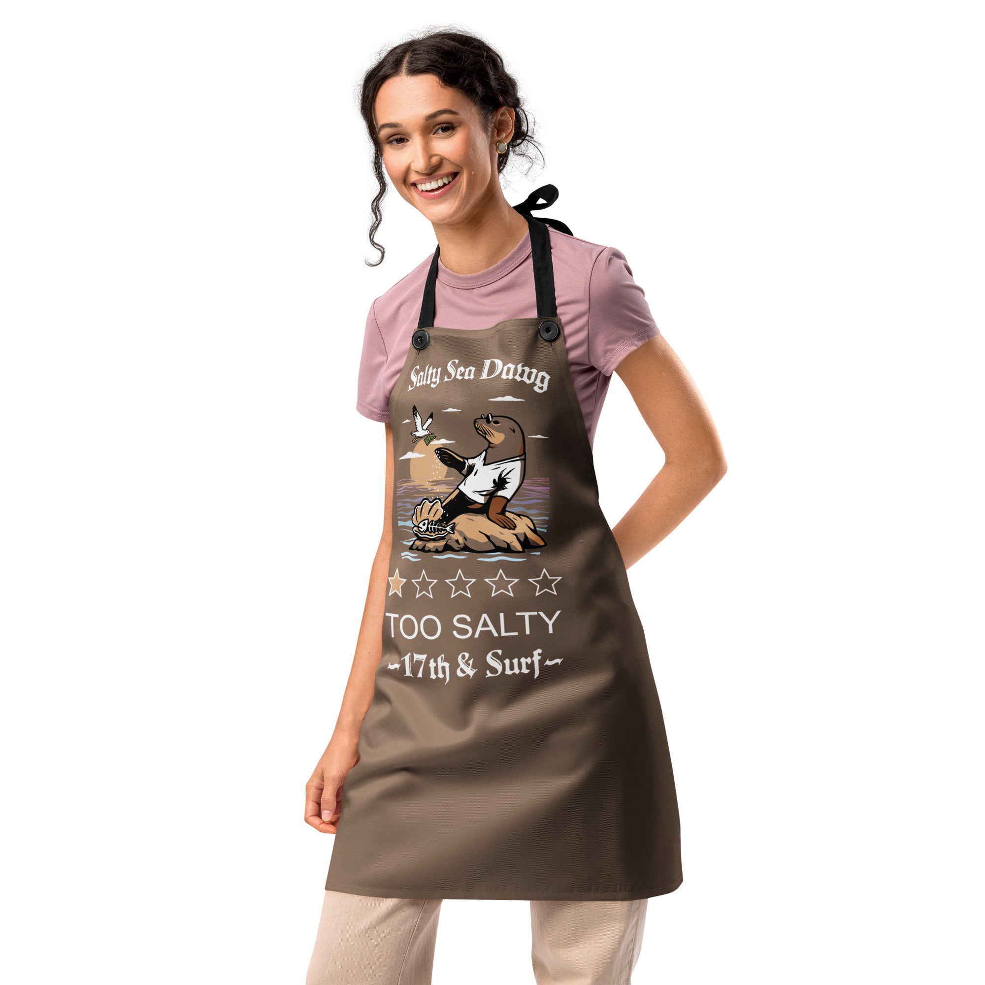 Model wearing "Salty Sea Dawg" chef apron featuring color graphic of a sea lion sprinkling salt on a clam shell with a fish skeleton beneath arched "Salty Sea Dawg" text and "TOO SALTY" one-star rating with "17th & Surf" wordmark, second front view – 17th & Surf