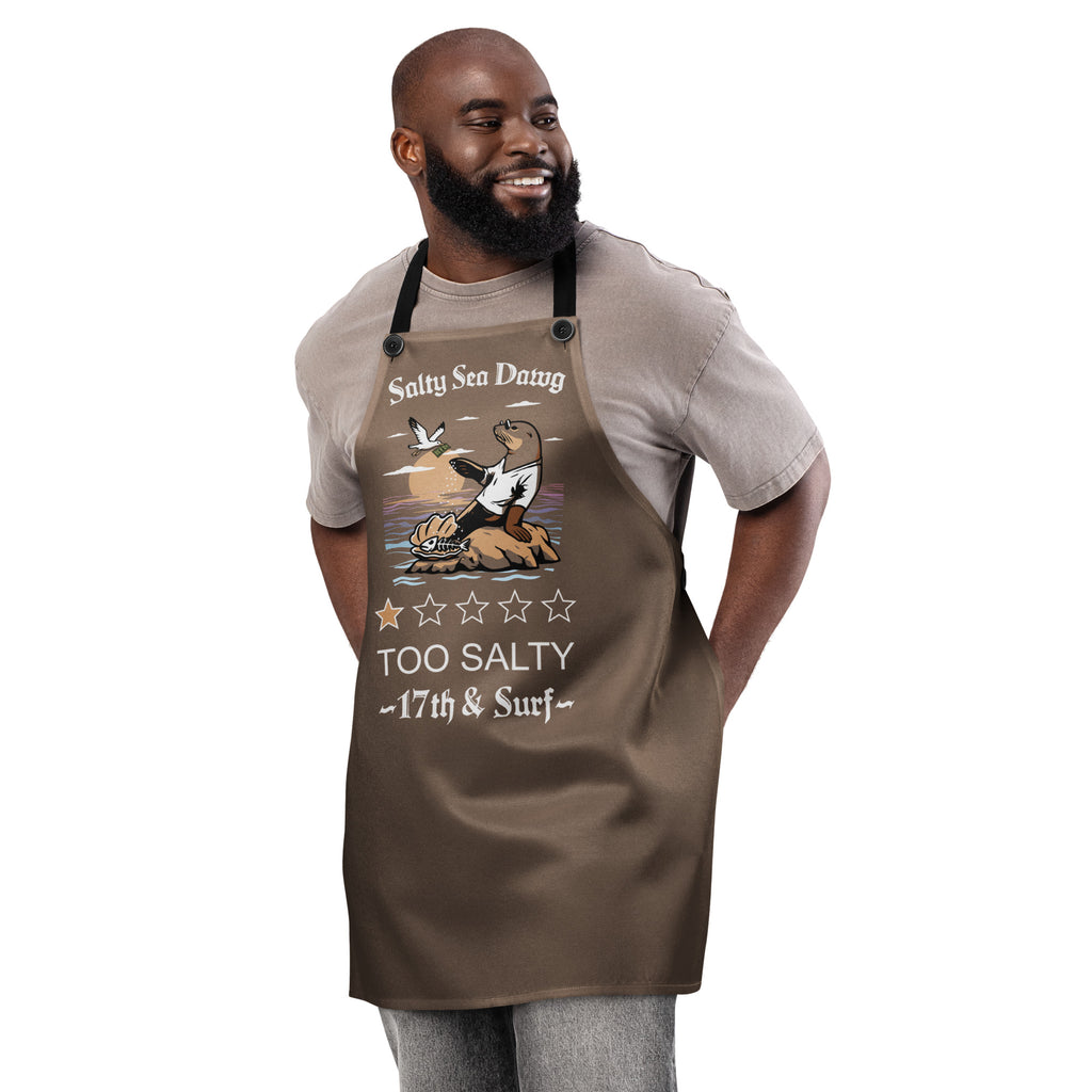 Model wearing "Salty Sea Dawg" chef apron featuring color graphic of a sea lion sprinkling salt on a clam shell with a fish skeleton beneath arched "Salty Sea Dawg" text and "TOO SALTY" one-star rating with "17th & Surf" wordmark, front view – 17th & Surf