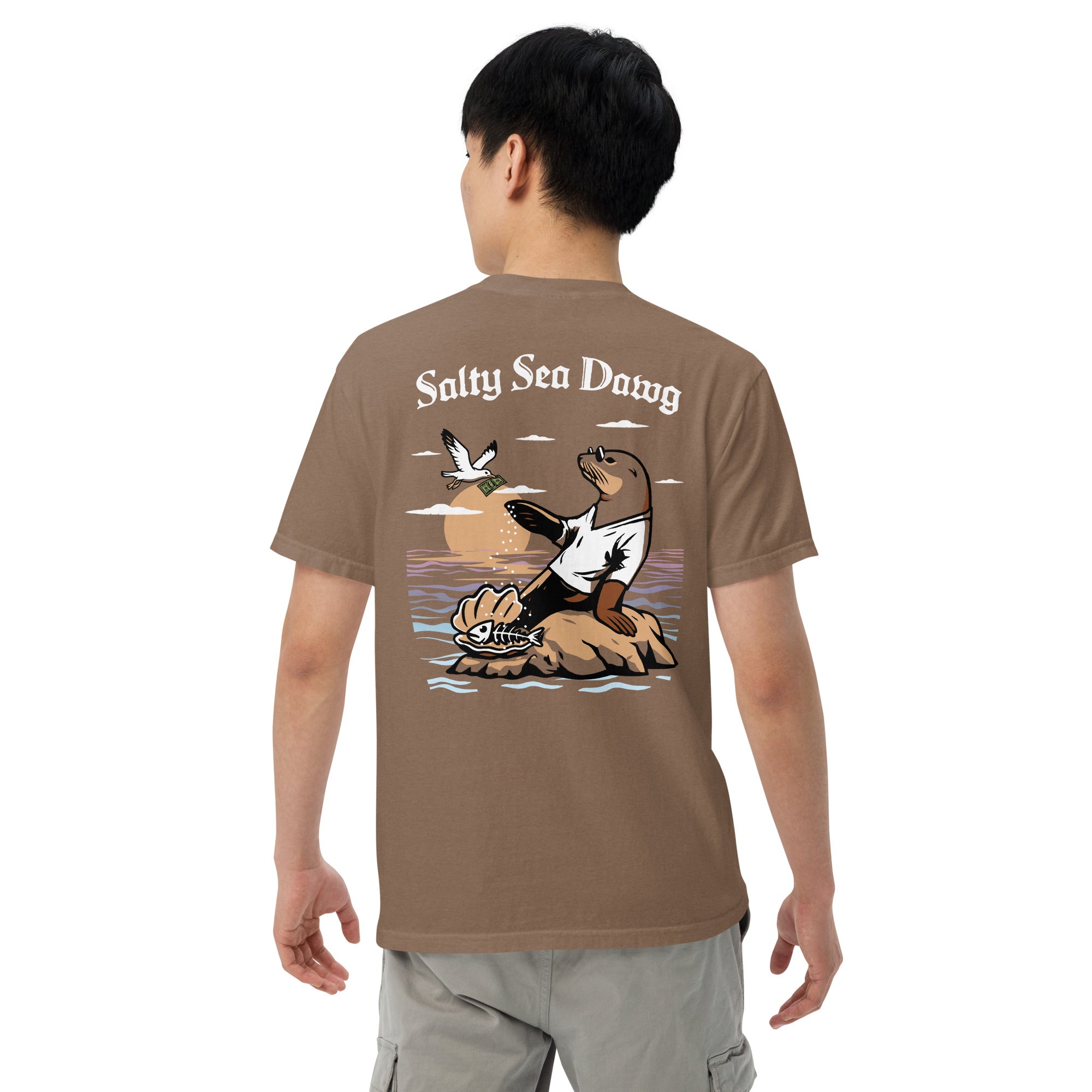 Model wearing "Salty Sea Dawg" garment-dyed heavyweight t-shirt in espresso, back angle view featuring color graphic of a sea lion sprinkling salt on a clam shell with a fish skeleton beneath arched "Salty Sea Dawg" text – 17th & Surf