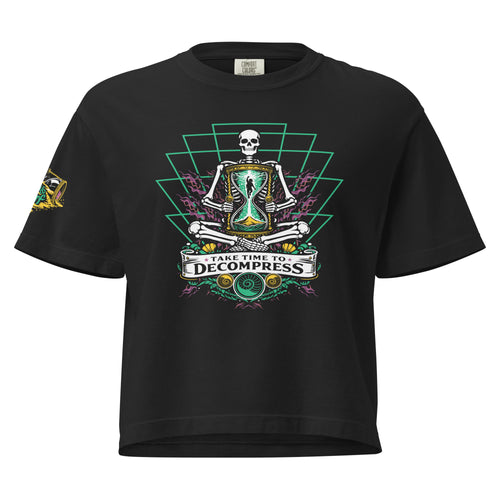 "Take Time To Decompress" black heavyweight boxy t-shirt, front flat lay, featuring a meditating skeleton holding a glowing hourglass with a tiny silhouette inside, framed by a green geometric pyramid pattern with purple coral accents and gold ammonite shells, above a "Take Time To Decompress" banner, with a small gold and teal "17th & Surf" hourglass logo on the sleeve – 17th & Surf