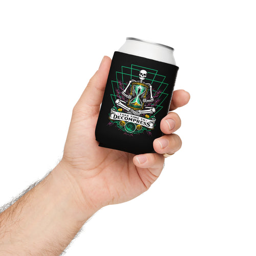 "Take Time To Decompress" black 12oz regular can cooler, held in hand, featuring a meditating skeleton holding a glowing hourglass with a tiny silhouette inside, framed by a green geometric pyramid pattern with purple coral accents and gold ammonite shells, above a "Take Time To Decompress" banner – 17th & Surf