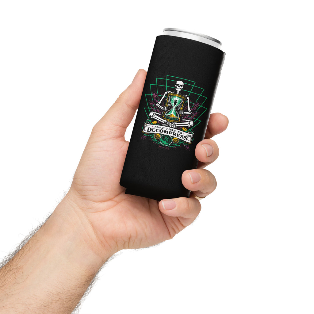 "Take Time To Decompress" black 12oz slim can cooler, held in hand, featuring a meditating skeleton holding a glowing hourglass with a tiny silhouette inside, framed by a green geometric pyramid pattern with purple coral accents and gold ammonite shells, above a "Take Time To Decompress" banner – 17th & Surf