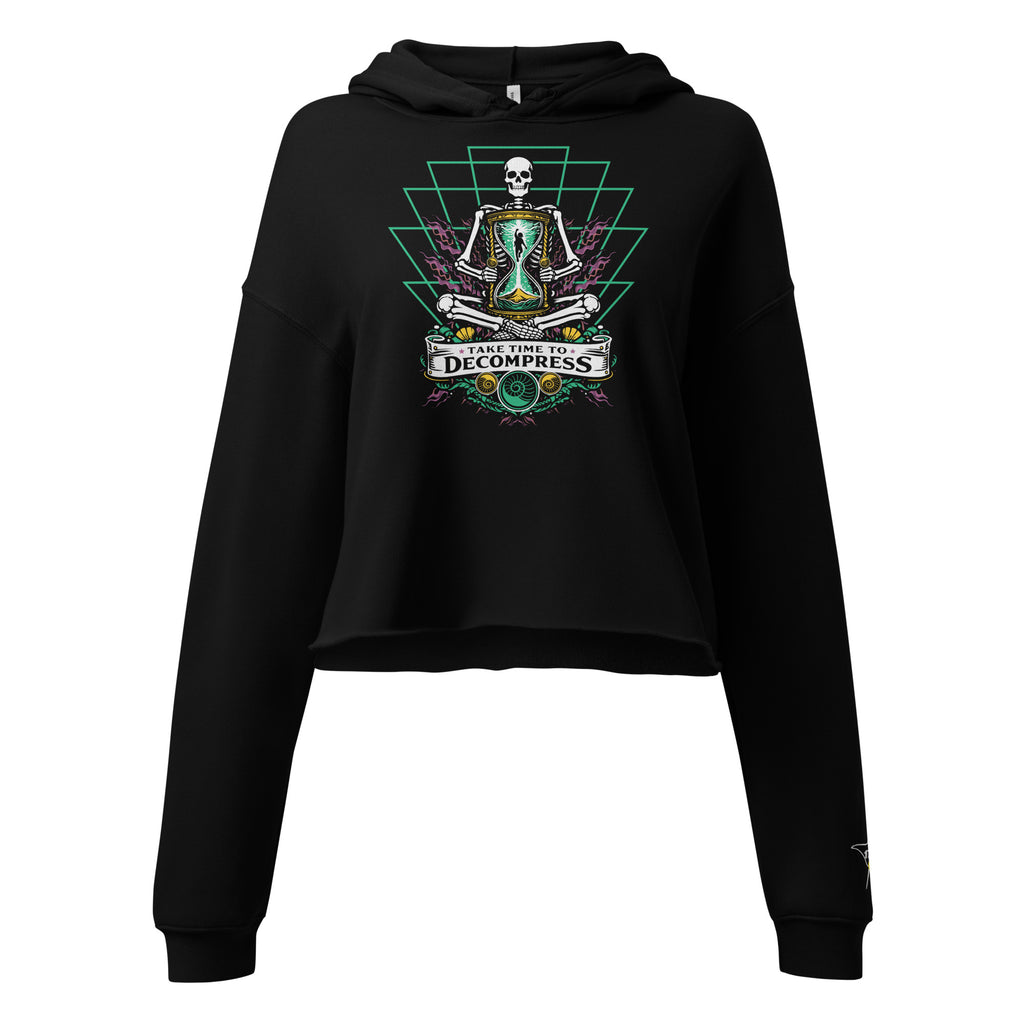"Take Time To Decompress" black cropped hoodie, front flat lay, featuring a meditating skeleton holding a glowing hourglass with a tiny silhouette inside, framed by a green geometric pyramid pattern with purple coral accents and gold ammonite shells, above a "Take Time To Decompress" banner – 17th & Surf