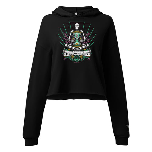 "Take Time To Decompress" black cropped hoodie, front flat lay, featuring a meditating skeleton holding a glowing hourglass with a tiny silhouette inside, framed by a green geometric pyramid pattern with purple coral accents and gold ammonite shells, above a "Take Time To Decompress" banner – 17th & Surf