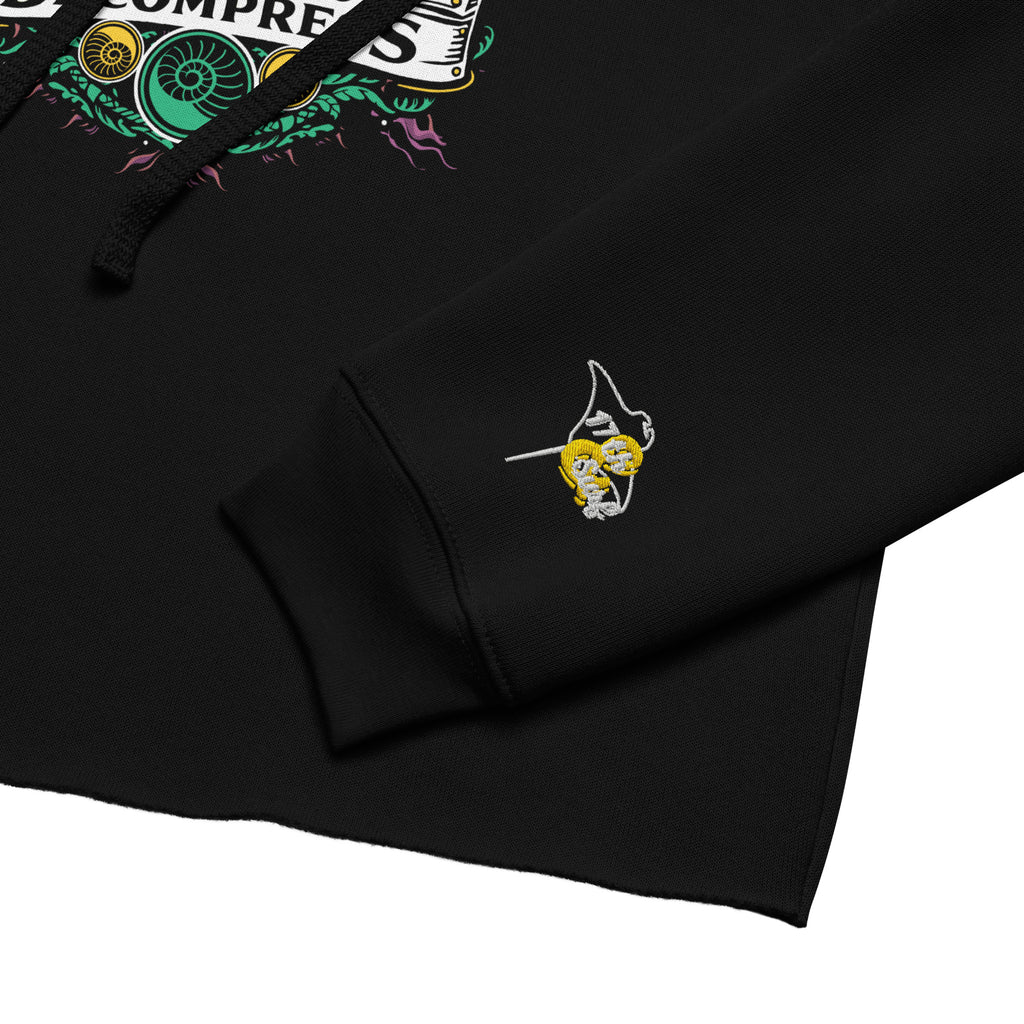 "Take Time To Decompress" black cropped hoodie, close-up of sleeve cuff featuring the embroidered gold and teal "17th & Surf" hourglass logo with a corner of the main meditating skeleton graphic visible – 17th & Surf
