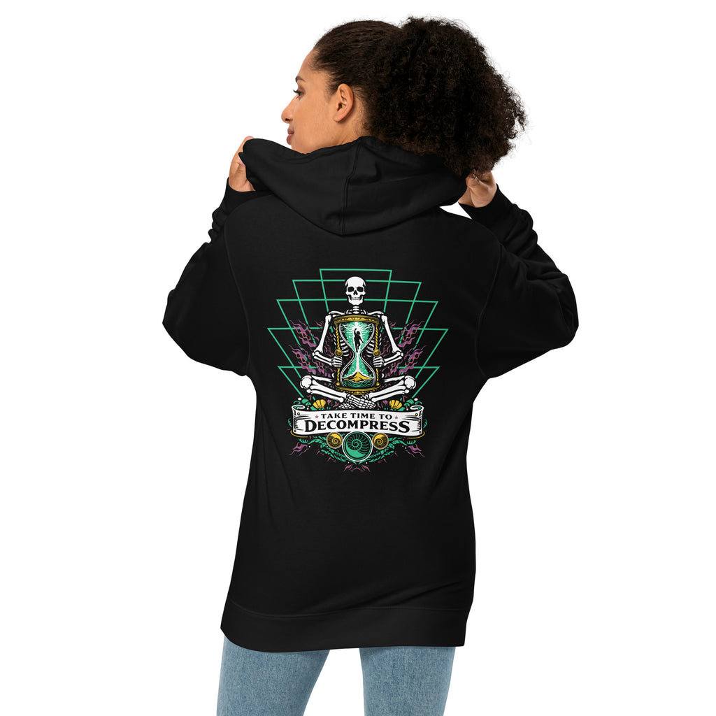 "Take Time To Decompress" black midweight hoodie on female model, back view, featuring a meditating skeleton holding a glowing hourglass with a tiny silhouette inside, framed by a green geometric pyramid pattern with purple coral accents and gold ammonite shells, above a "Take Time To Decompress" banner – 17th & Surf