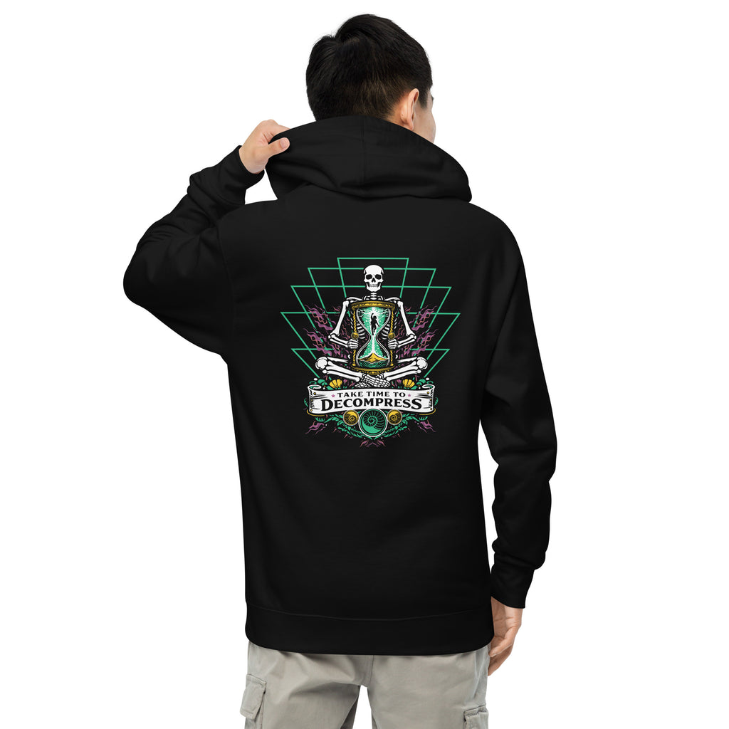 "Take Time To Decompress" black midweight hoodie on male model, back view, featuring a meditating skeleton holding a glowing hourglass with a tiny silhouette inside, framed by a green geometric pyramid pattern with purple coral accents and gold ammonite shells, above a "Take Time To Decompress" banner – 17th & Surf