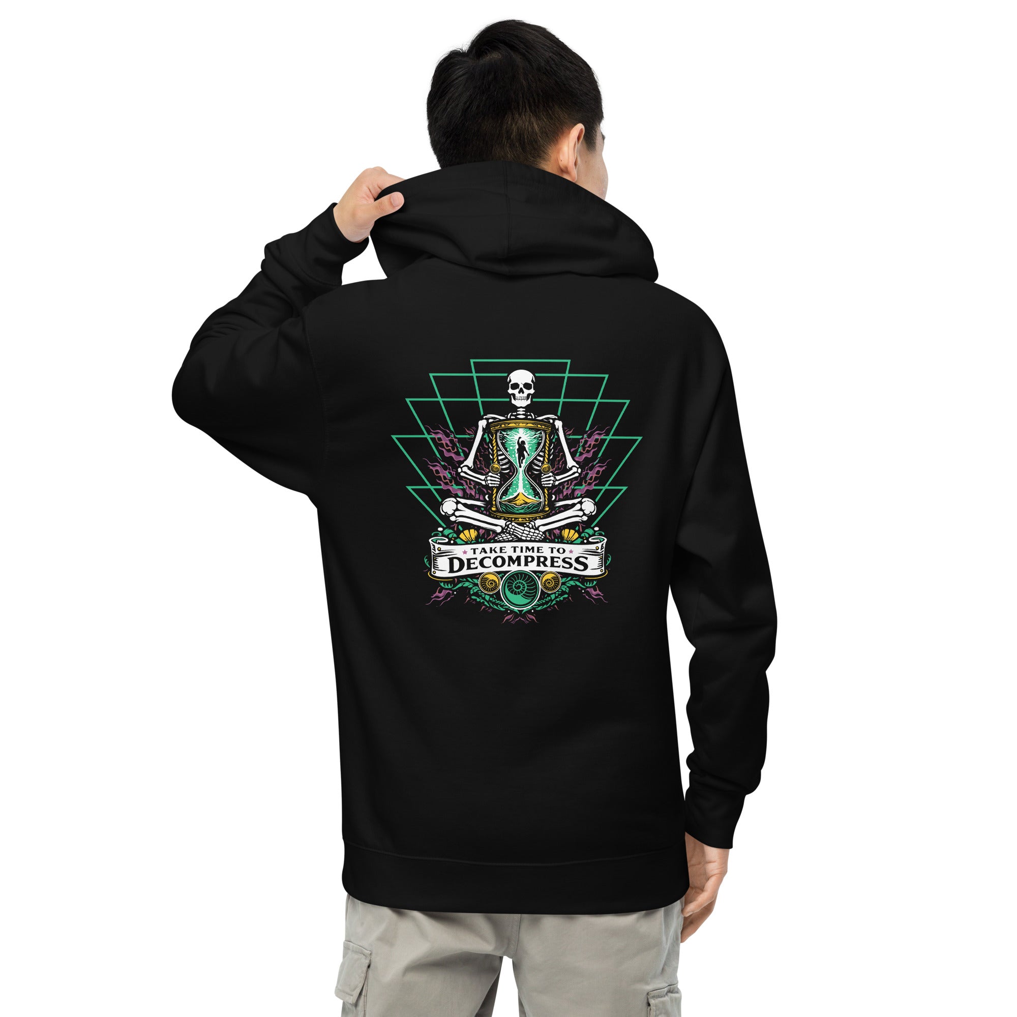 "Take Time To Decompress" black midweight hoodie on male model, back view, featuring a meditating skeleton holding a glowing hourglass with a tiny silhouette inside, framed by a green geometric pyramid pattern with purple coral accents and gold ammonite shells, above a "Take Time To Decompress" banner – 17th & Surf