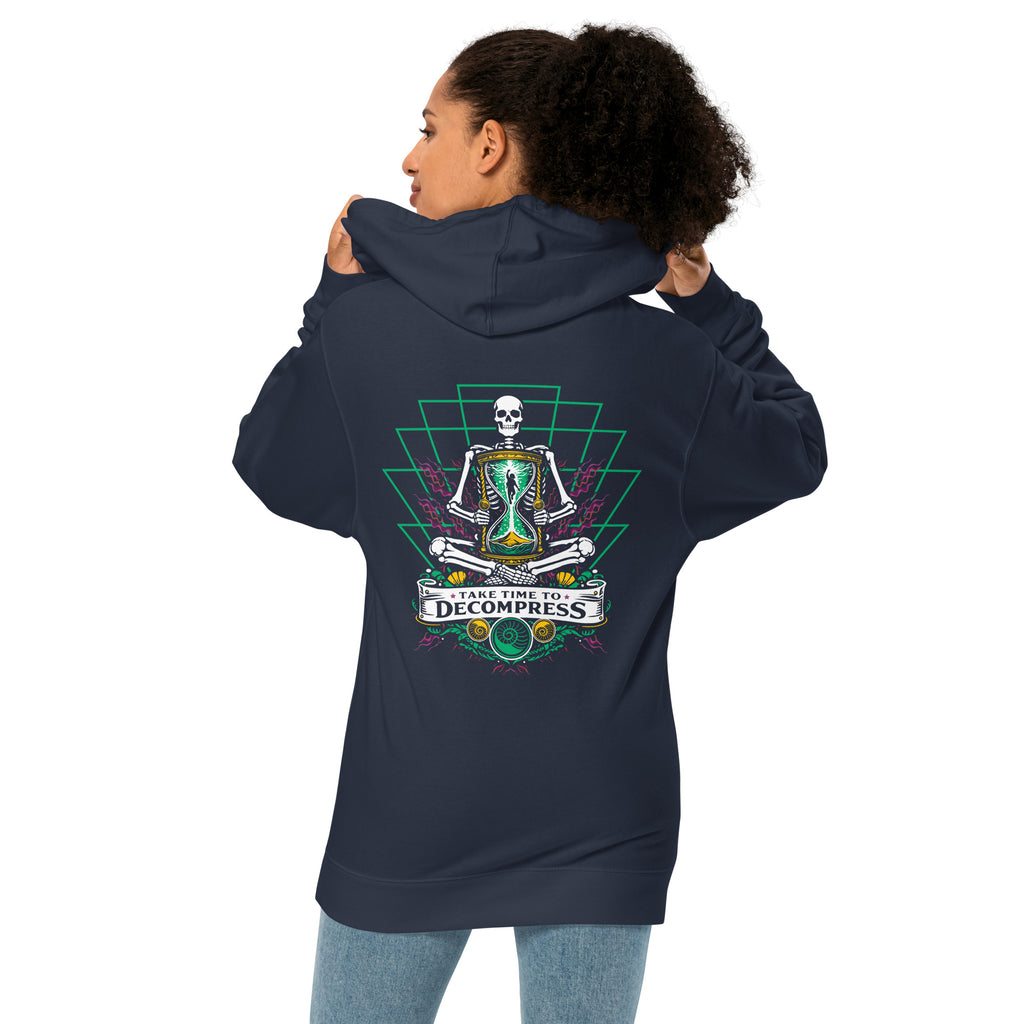 "Take Time To Decompress" classic navy midweight hoodie on female model, back view, featuring a meditating skeleton holding a glowing hourglass with a tiny silhouette inside, framed by a green geometric pyramid pattern with purple coral accents and gold ammonite shells, above a "Take Time To Decompress" banner – 17th & Surf