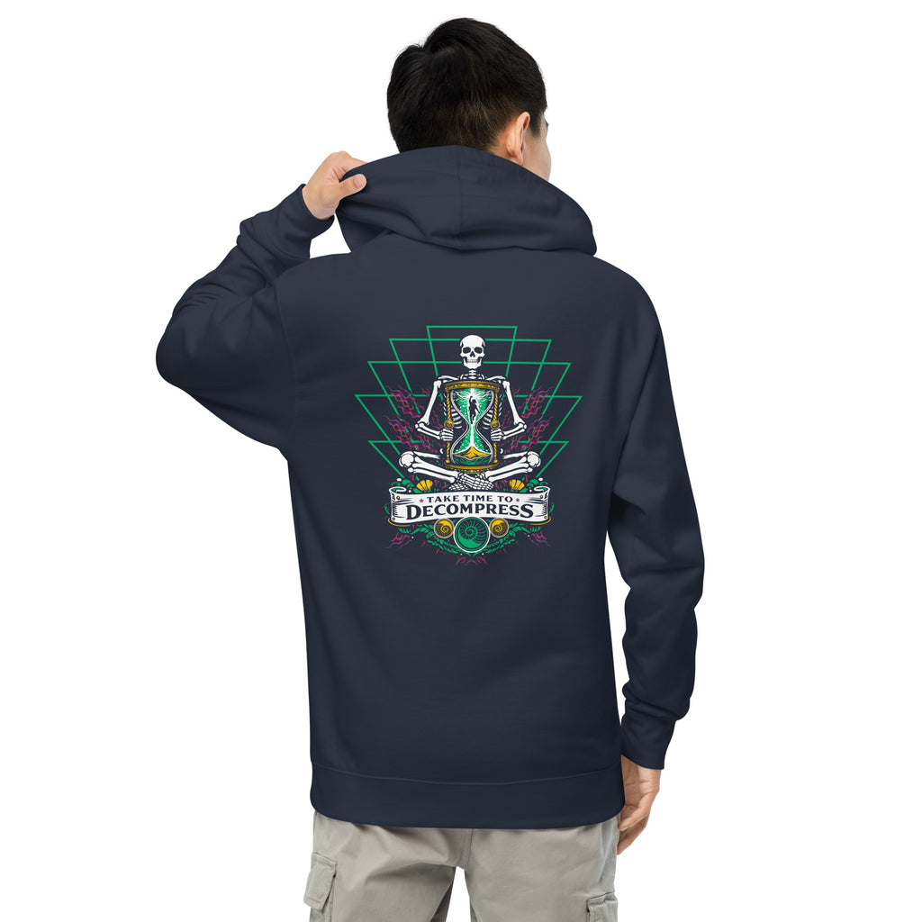 "Take Time To Decompress" classic navy midweight hoodie on male model, back view, featuring a meditating skeleton holding a glowing hourglass with a tiny silhouette inside, framed by a green geometric pyramid pattern with purple coral accents and gold ammonite shells, above a "Take Time To Decompress" banner – 17th & Surf