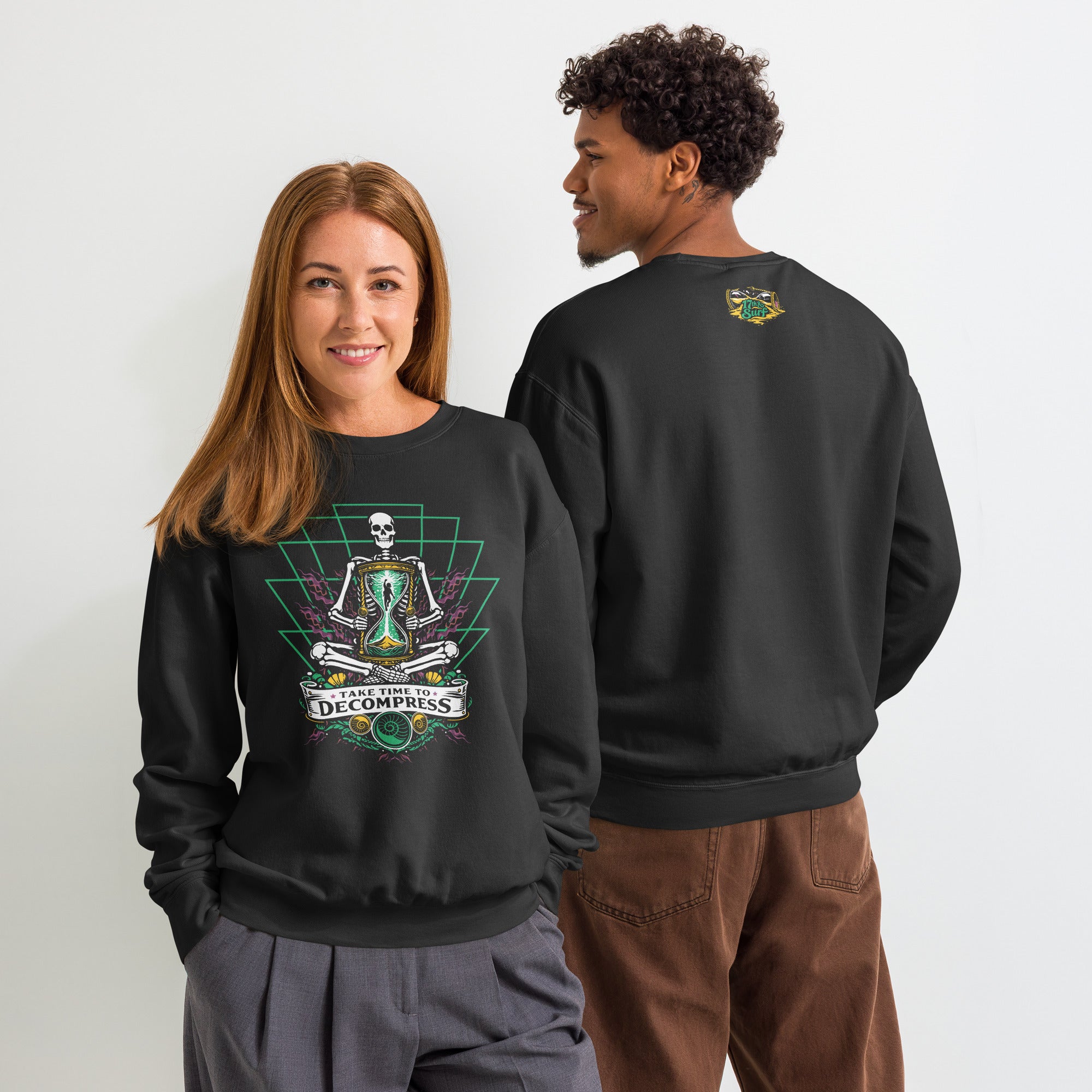 "Take Time To Decompress" black lightweight crewneck sweatshirt on two models, front view showing the full meditating skeleton design with glowing hourglass and back view showing a small gold and teal "17th & Surf" hourglass logo near the collar – 17th & Surf