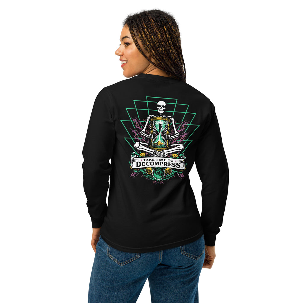 "Take Time To Decompress" black heavyweight long sleeve shirt on female model, back view, featuring a meditating skeleton holding a glowing hourglass with a tiny silhouette inside, framed by a green geometric pyramid pattern with purple coral accents and gold ammonite shells, above a "Take Time To Decompress" banner – 17th & Surf