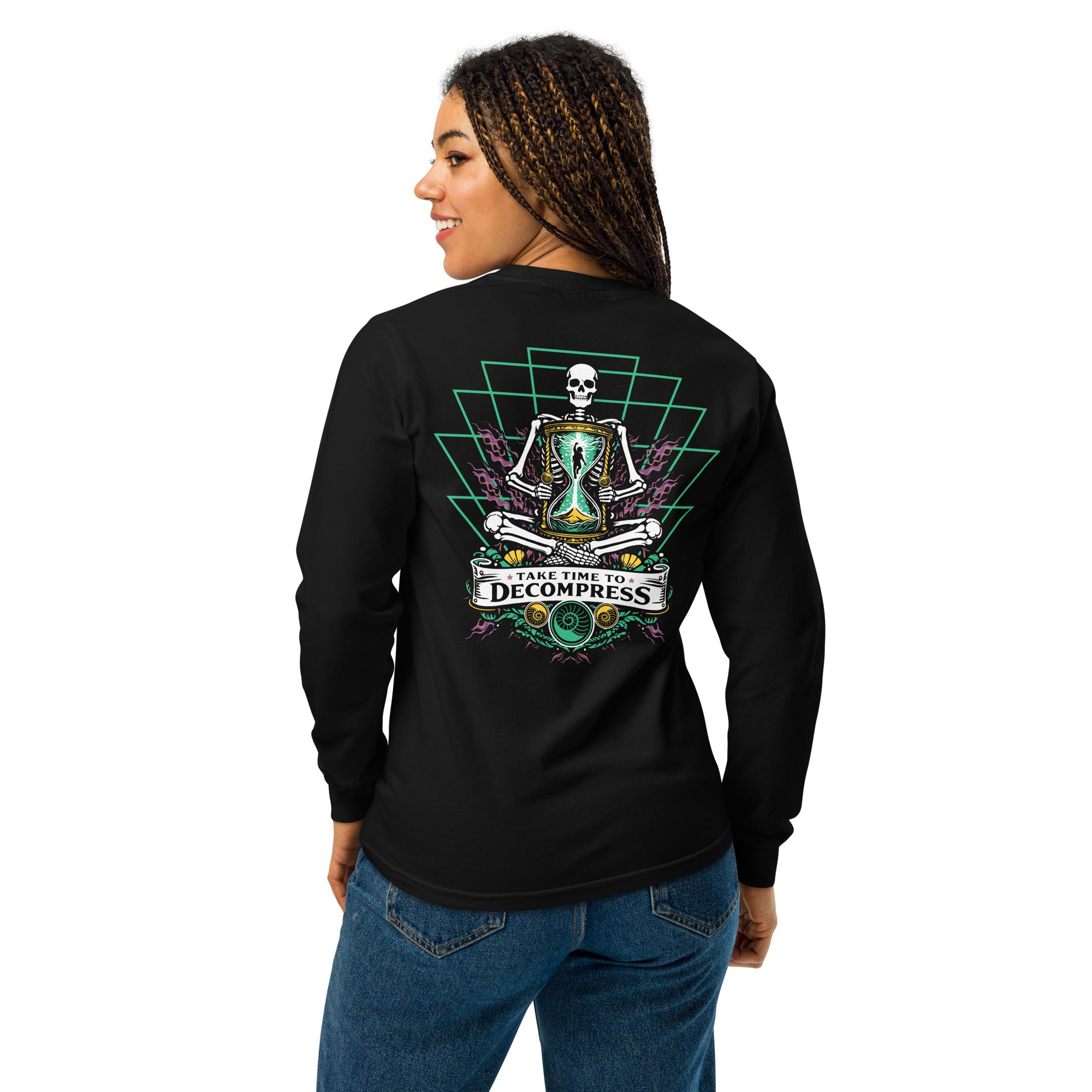 "Take Time To Decompress" black heavyweight long sleeve shirt on female model, back view, featuring a meditating skeleton holding a glowing hourglass with a tiny silhouette inside, framed by a green geometric pyramid pattern with purple coral accents and gold ammonite shells, above a "Take Time To Decompress" banner – 17th & Surf
