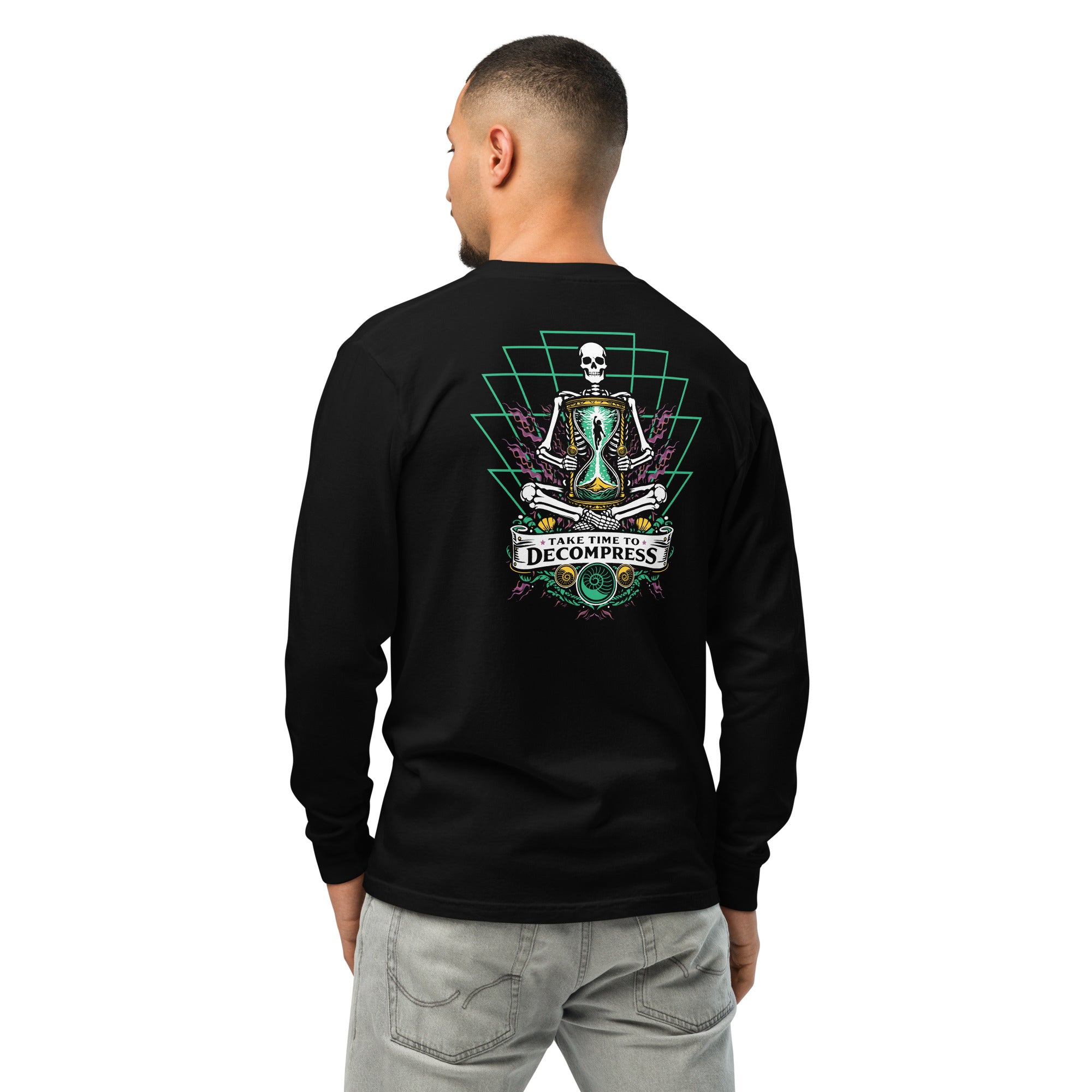 "Take Time To Decompress" black heavyweight long sleeve shirt on male model, back view, featuring a meditating skeleton holding a glowing hourglass with a tiny silhouette inside, framed by a green geometric pyramid pattern with purple coral accents and gold ammonite shells, above a "Take Time To Decompress" banner – 17th & Surf