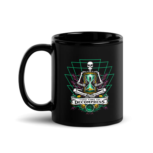 "Take Time To Decompress" black glossy 11oz mug, handle on left, featuring a meditating skeleton holding a glowing hourglass with a tiny silhouette inside, framed by a green geometric pyramid pattern with purple coral accents and gold ammonite shells, above a "Take Time To Decompress" banner – 17th & Surf