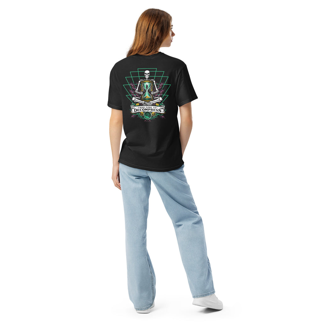 "Take Time To Decompress" black heavyweight t-shirt on female model, back view, featuring a meditating skeleton holding a glowing hourglass with a tiny silhouette inside, framed by a green geometric pyramid pattern with purple coral accents and gold ammonite shells, above a "Take Time To Decompress" banner – 17th & Surf