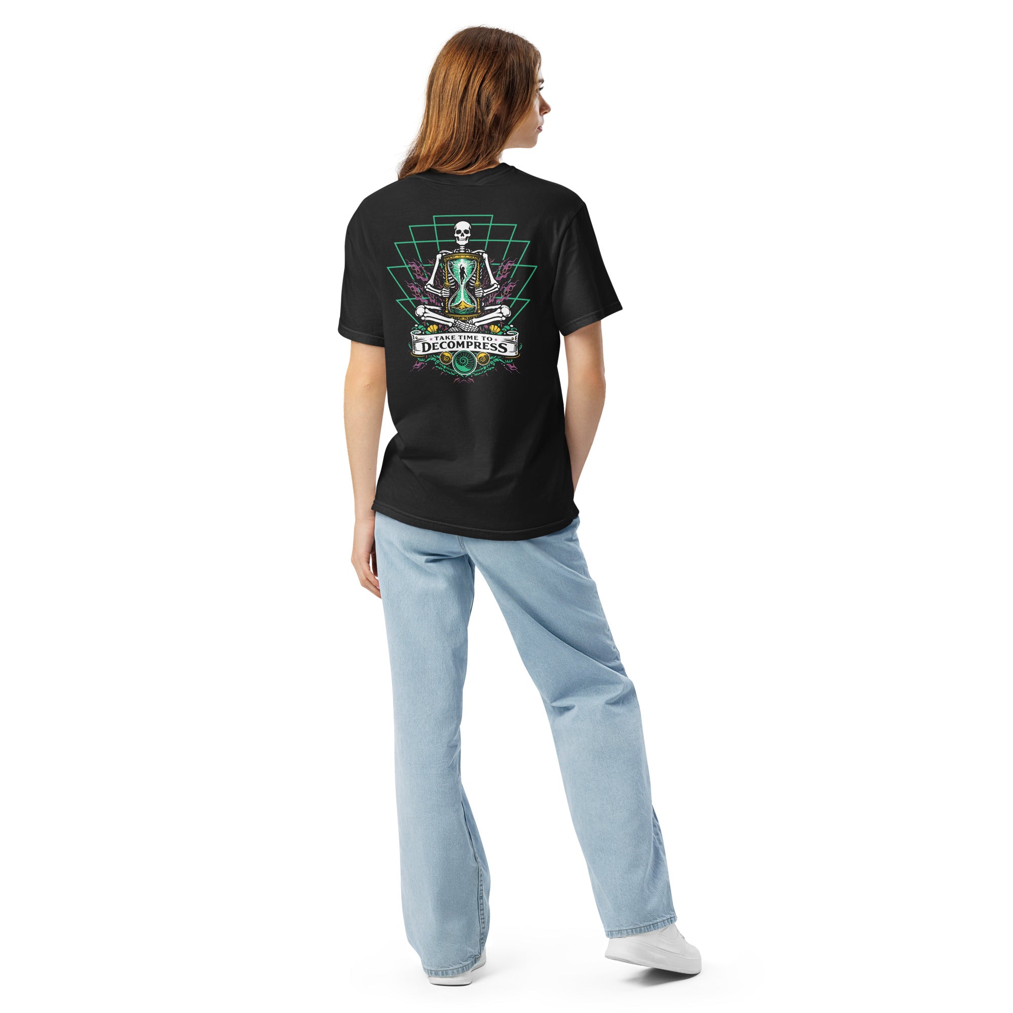 "Take Time To Decompress" black heavyweight t-shirt on female model, back view, featuring a meditating skeleton holding a glowing hourglass with a tiny silhouette inside, framed by a green geometric pyramid pattern with purple coral accents and gold ammonite shells, above a "Take Time To Decompress" banner – 17th & Surf