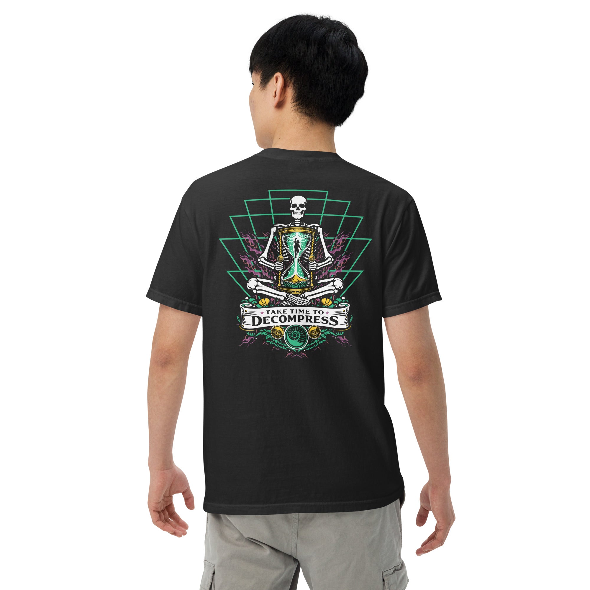 "Take Time To Decompress" black heavyweight t-shirt on male model, back view, featuring a meditating skeleton holding a glowing hourglass with a tiny silhouette inside, framed by a green geometric pyramid pattern with purple coral accents and gold ammonite shells, above a "Take Time To Decompress" banner – 17th & Surf
