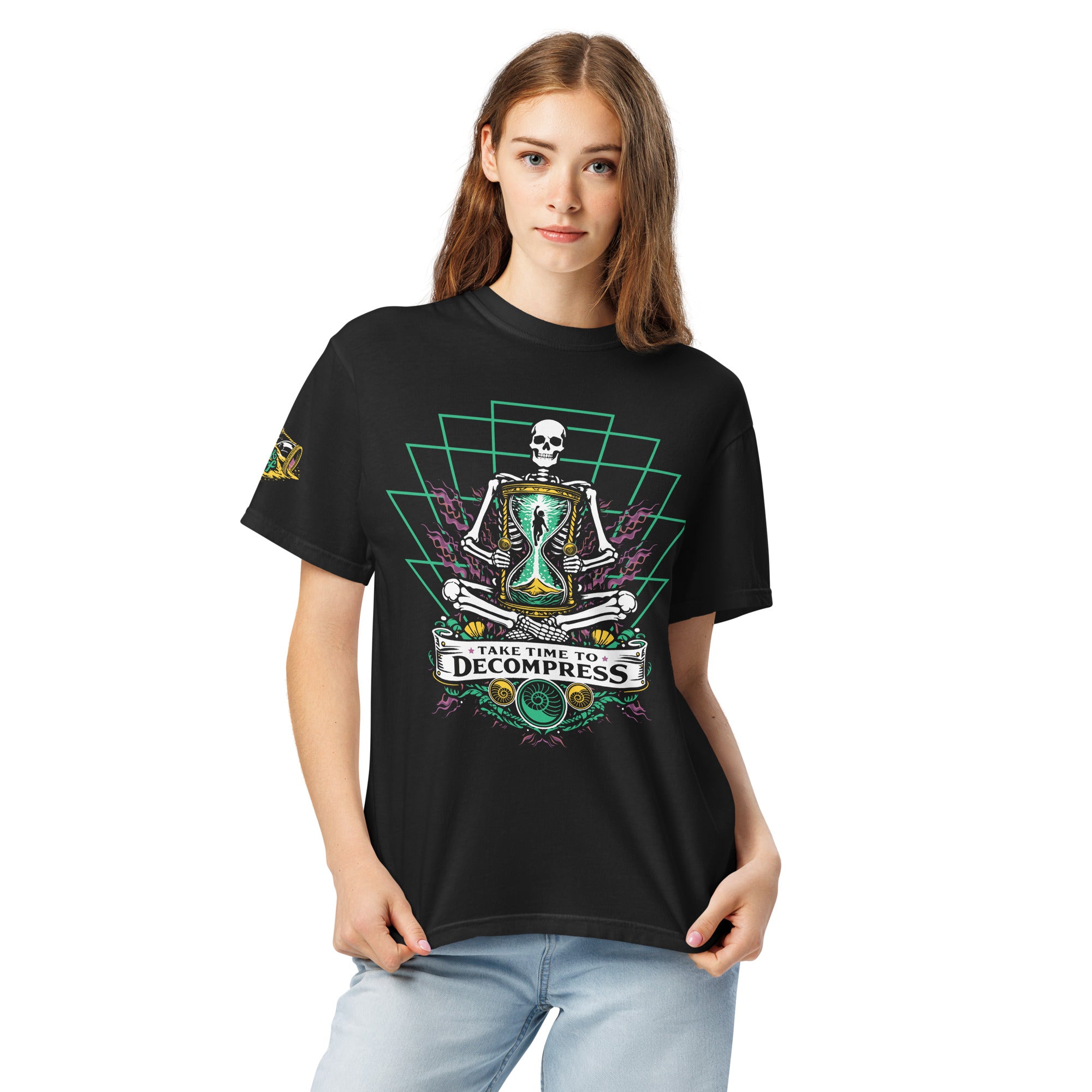 "Take Time To Decompress" black heavyweight t-shirt on female model, front view, featuring a meditating skeleton holding a glowing hourglass with a tiny silhouette inside, framed by a green geometric pyramid pattern with purple coral accents and gold ammonite shells, above a "Take Time To Decompress" banner – 17th & Surf