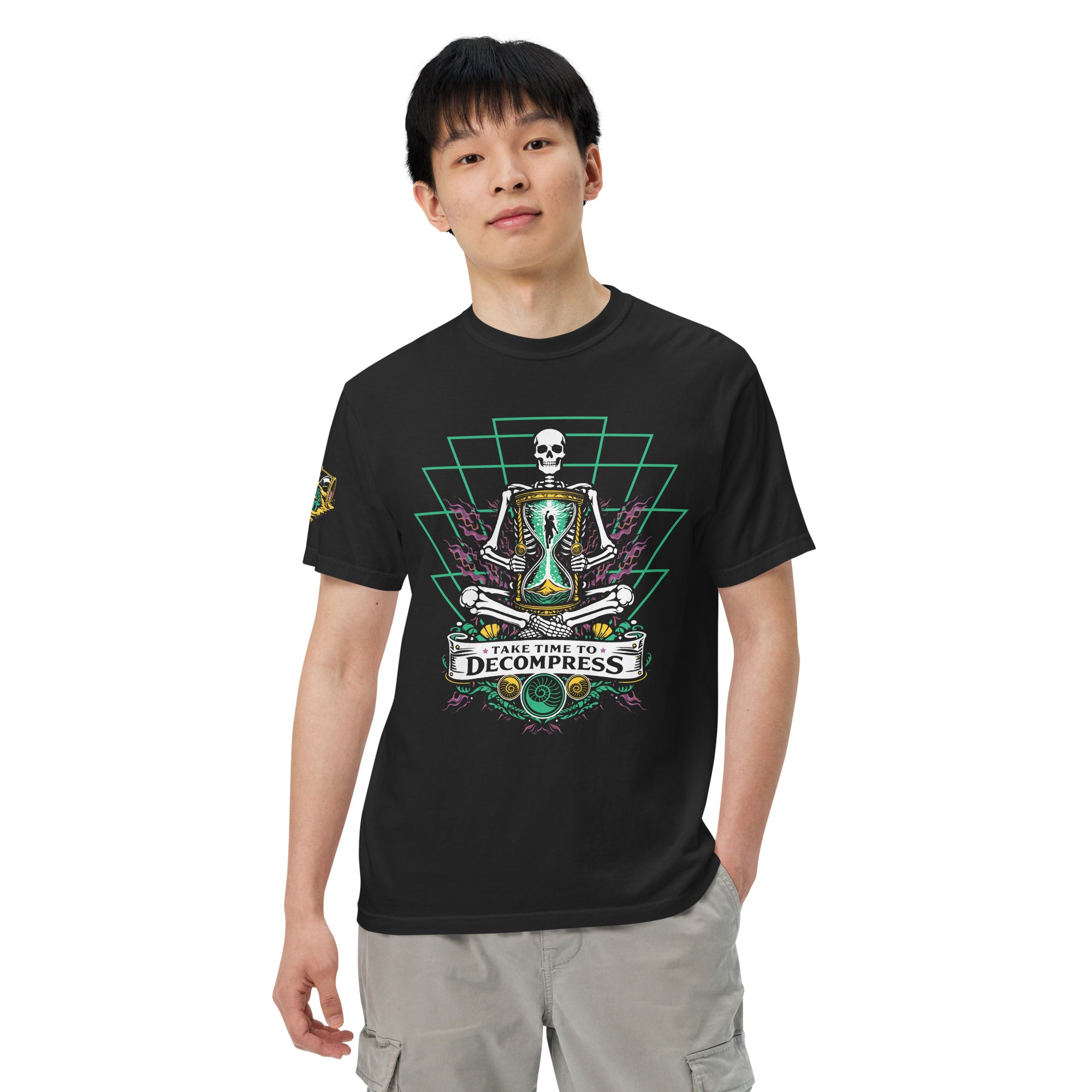 "Take Time To Decompress" black heavyweight t-shirt on male model, front view, featuring a meditating skeleton holding a glowing hourglass with a tiny silhouette inside, framed by a green geometric pyramid pattern with purple coral accents and gold ammonite shells, above a "Take Time To Decompress" banner – 17th & Surf